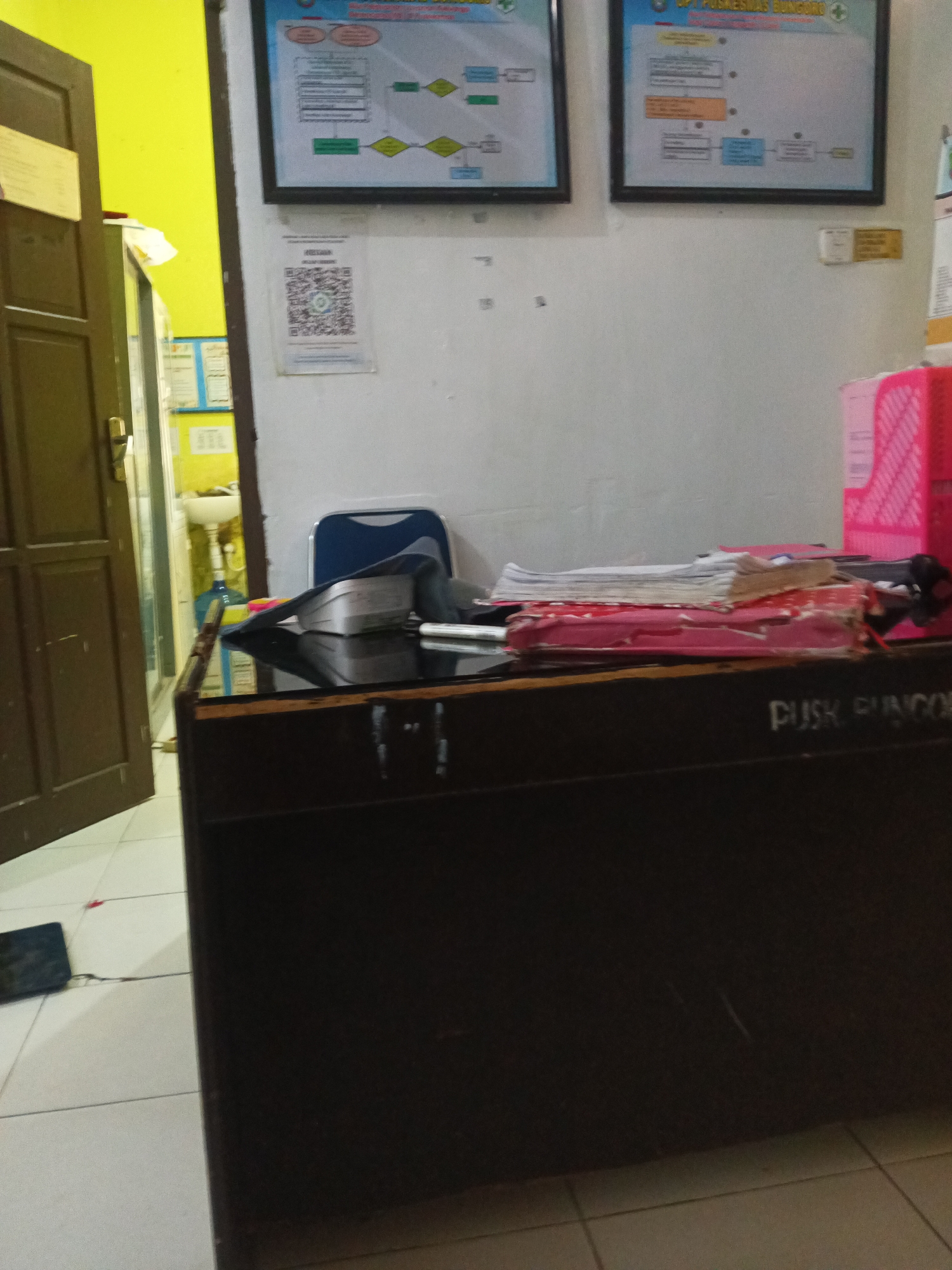 The image depicts an interior scene, likely an office or administrative area, in Paddoang Doangan, Indonesia. The dominant features are a dark-colored desk in the foreground, laden with stacks of papers and folders. A blue and black chair is partially visible behind the desk. To the left, a partially opened brown wooden door reveals a bright yellow wall and a glimpse of a sink and water dispenser in an adjacent room. On the wall behind the desk, two framed charts or diagrams are displayed, along with a QR code mounted on the wall. To the right, a pink plastic organizer holds additional pink items. The floor is tiled with white and beige squares. The overall impression is one of a functional, if somewhat cluttered, workspace.