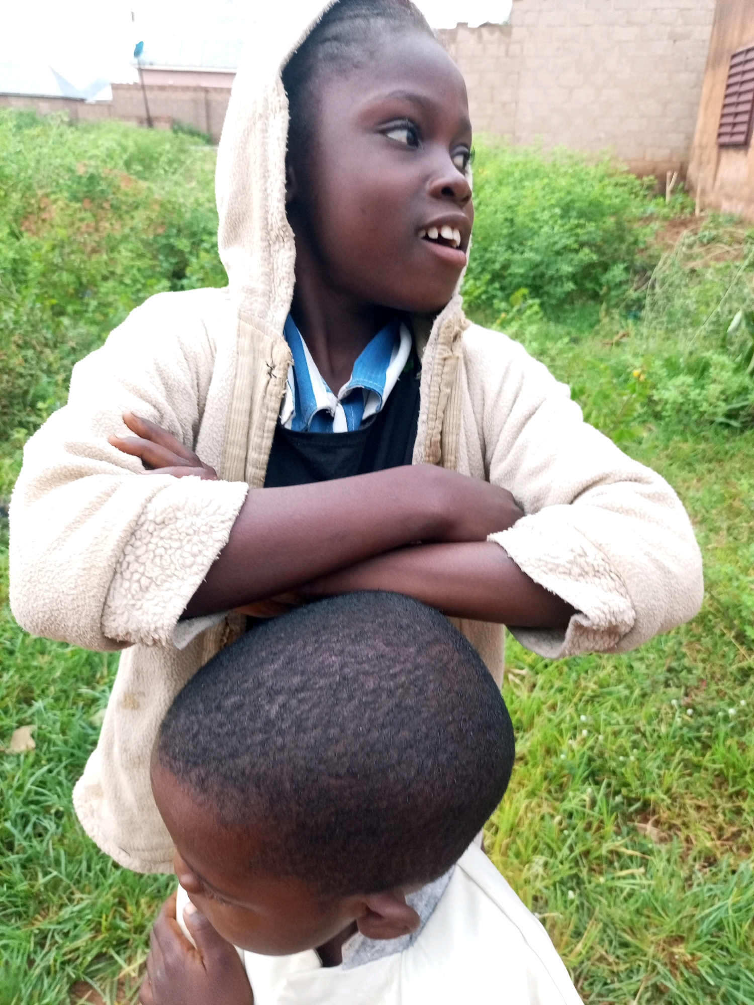 Two children are in a grassy outdoor area in Tempure, Nigeria. The child in the foreground is facing away from the camera, with their head bent down. Their dark hair is short and closely cropped. They are wearing a light-colored garment. The child behind them is facing to the right, with their arms crossed. They are wearing a cream-colored hooded jacket over a blue and white striped shirt. The background consists of green grass and some vegetation, with buildings and fences visible in the distance.