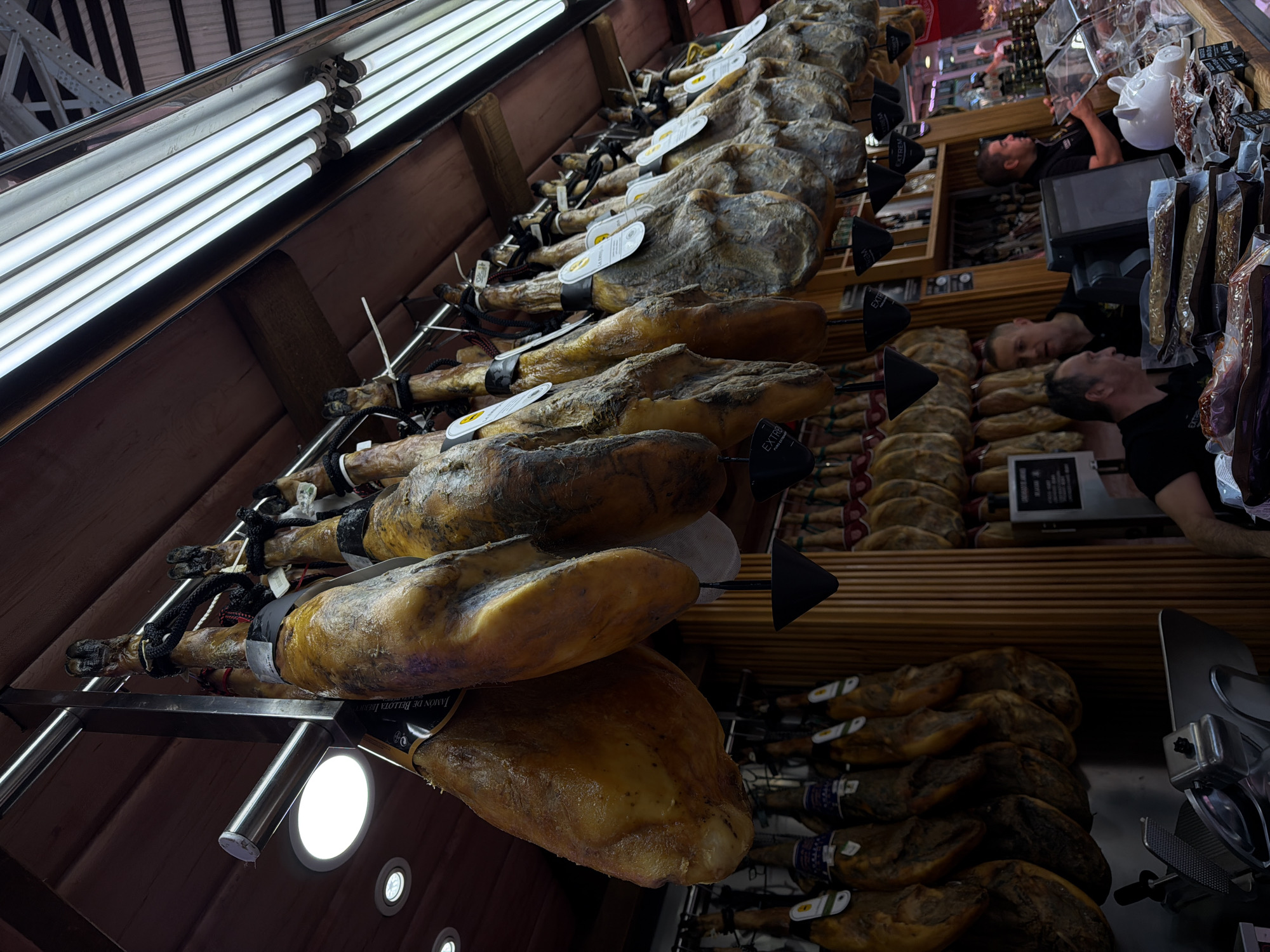 This photograph depicts a butcher shop in the city of València, Spain. Multiple cured ham legs are suspended from a metal rack in the foreground. Two butchers, visible in the background, are positioned behind a counter where processed hams are displayed. The hams are arranged in an orderly fashion along the shelves. The lighting is provided by fluorescent ceiling lights above the ham legs.