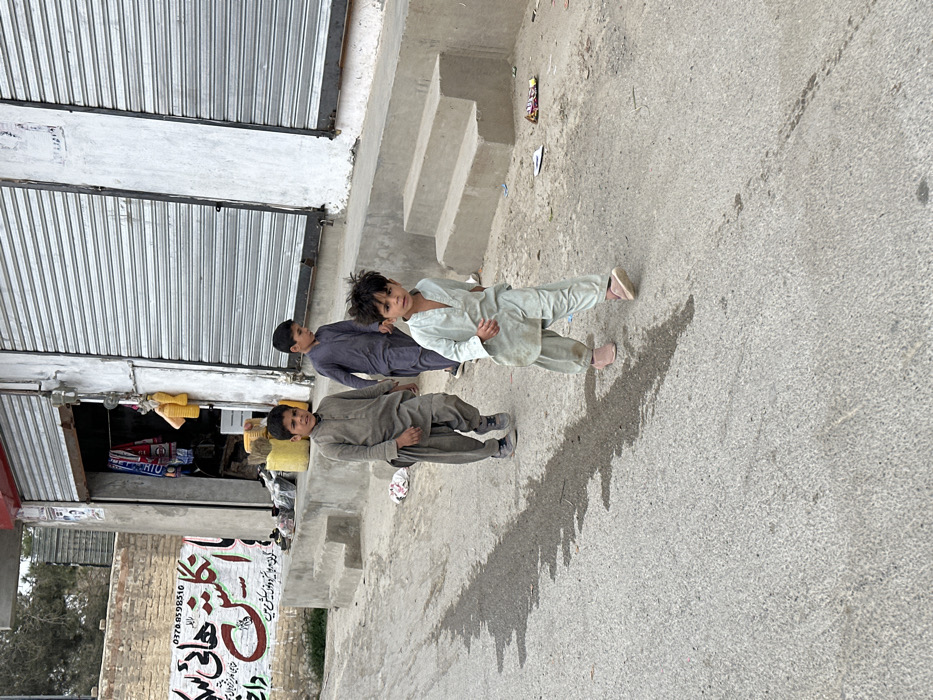 This image depicts an outdoor urban scene in Quetta City Tehsil, Pakistan. Three boys are walking on an asphalt road. The youngest boy, estimated to be 3-5 years old, is in the foreground, wearing a pale green shalwar kameez and pink shoes, looking towards the camera. Behind him, and slightly to the left, is a boy approximately 6-8 years old, dressed in a grey-brown shalwar kameez and dark shoes, also facing the camera. The third boy, positioned further back and to the left, estimated to be 7-9 years old, wears a dark blue/purple shalwar kameez and dark shoes, looking forward.
To the right, a building with light-colored walls features two closed, silver-colored metal roll-down shutters. An open doorway, located between the shutters and slightly to the left, reveals a dimly lit interior with hanging items and yellow containers visible. Concrete steps ascend from the road to the building's entrance level. To the left, a brick wall displays a large, red-painted sign with script (likely Urdu or Pashto) and a phone number (03708598516), with smaller white script below. A dark, irregular patch, consistent with a wet area or a distinct shadow, is present on the asphalt road surface. Scattered litter, including wrappers, is visible on the ground near the building steps.
