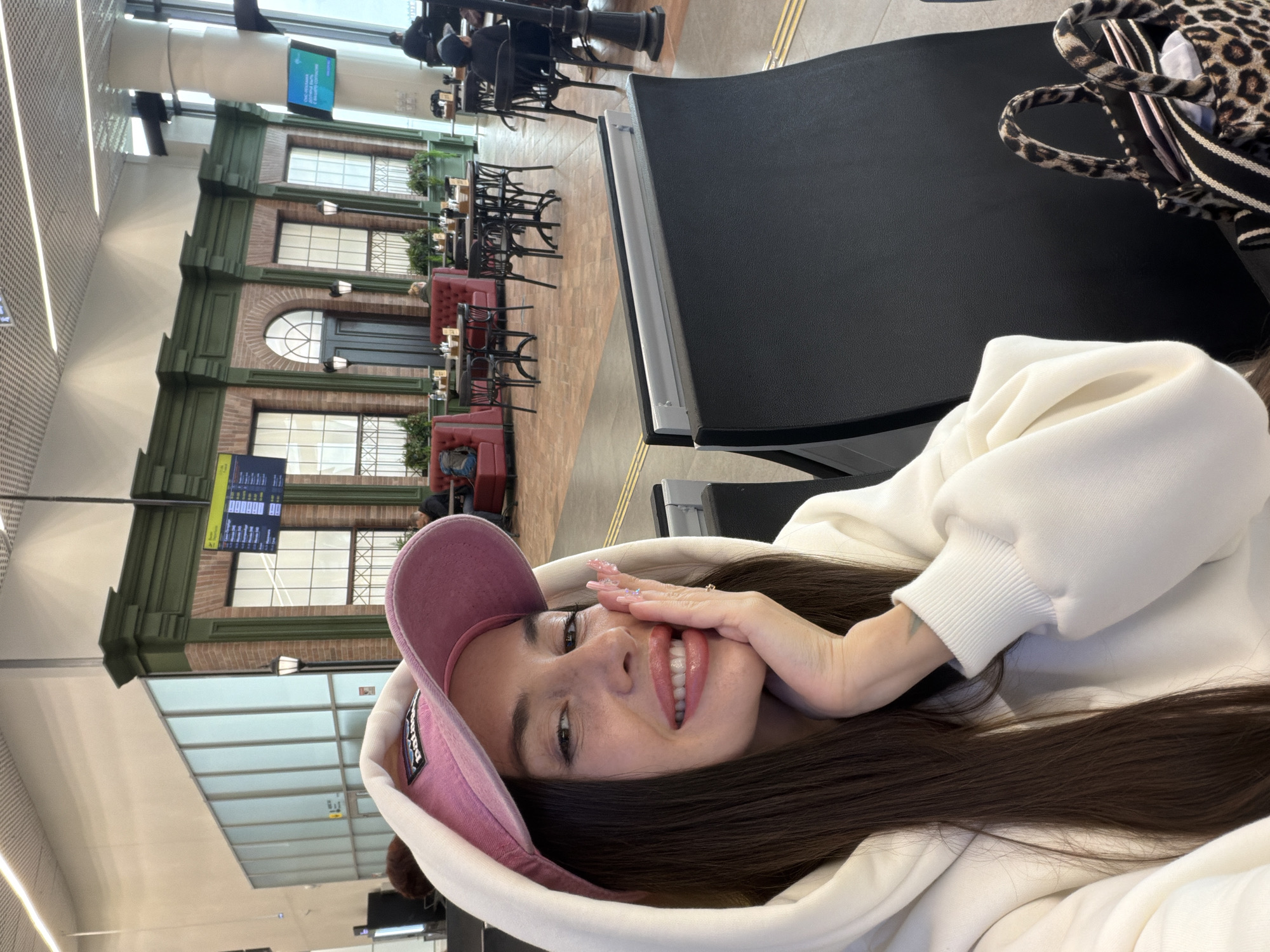 The image, originally rotated 90 degrees clockwise, captures a young woman taking a selfie in what appears to be a large, modern indoor public space, likely an airport terminal in Новое Мартьяново, Russia.
**Subjects:** The primary subject is a young woman, likely in her late teens or early twenties, with long, dark brown hair. She is smiling broadly, showing her teeth, and has one hand covering part of her mouth, revealing long, manicured nails with glitter. She wears a light-colored (possibly white or cream) hooded sweatshirt and a pink baseball cap with a visible "Patagonia" logo. In the background, several other indistinct people are seated at tables.
**Scene and Setting:** The setting is an expansive indoor area featuring a multi-story facade designed to resemble an old-fashioned street with brickwork, green trim, large windows (some with decorative grates), and an arched doorway. This facade creates the impression of an outdoor cafe within an indoor environment. There are numerous tables with red booth seating and black, ornate chairs. Overhead, large ventilation ducts and modern lighting fixtures are visible. The floor is tiled with a tactile yellow strip.
**Activity:** The woman is actively taking a selfie, holding the camera directly in front of her face. People in the background are seated, suggesting they are waiting, dining, or relaxing.
**Notable Details:** The presence of large digital display screens showing flight or general public information (text not clearly legible but appears to be in a foreign language) strongly indicates an airport or a major transportation hub. The woman appears happy and relaxed, directly engaging with the camera. A leopard-print tote bag is visible next to her. The perspective is a close-up selfie with a wide-angle view of the background.
