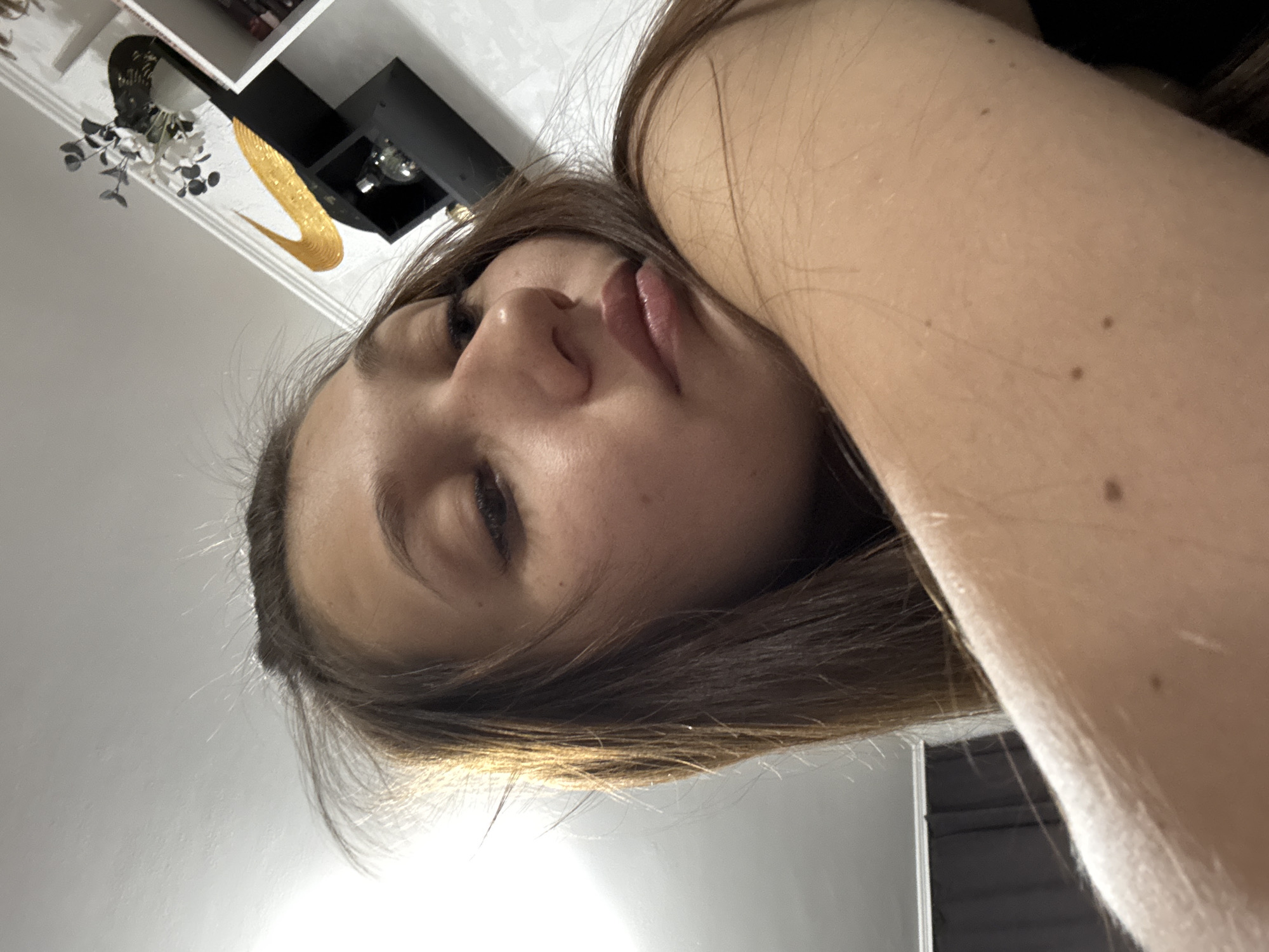 The media file is a close-up, low-angle selfie of a young woman lying down. Her face is the primary subject, tilted upwards and to the side, with her eyes looking towards the camera. Her hair, a shade of brown with lighter highlights, is spread around her head and shoulders. Her lips are slightly parted. A portion of her arm and shoulder are also visible in the foreground, showing some light freckles.
In the background, a shelf unit is visible on a white wall, with various objects placed on it. One object appears to be a black shelf with a shiny, golden, curved decoration resembling a swoosh, and a small, faceted glass object. Another shelf above it holds what looks like a bouquet of artificial white flowers with dark leaves. The overall lighting suggests an indoor setting, possibly a home, with the light source illuminating the scene from the side. The angle and lighting create an intimate and slightly abstract feel to the image. The context provided indicates this was taken in Novokuznetsk, Russia. No specific time of day or weather can be determined from the image itself, and no text is visible.