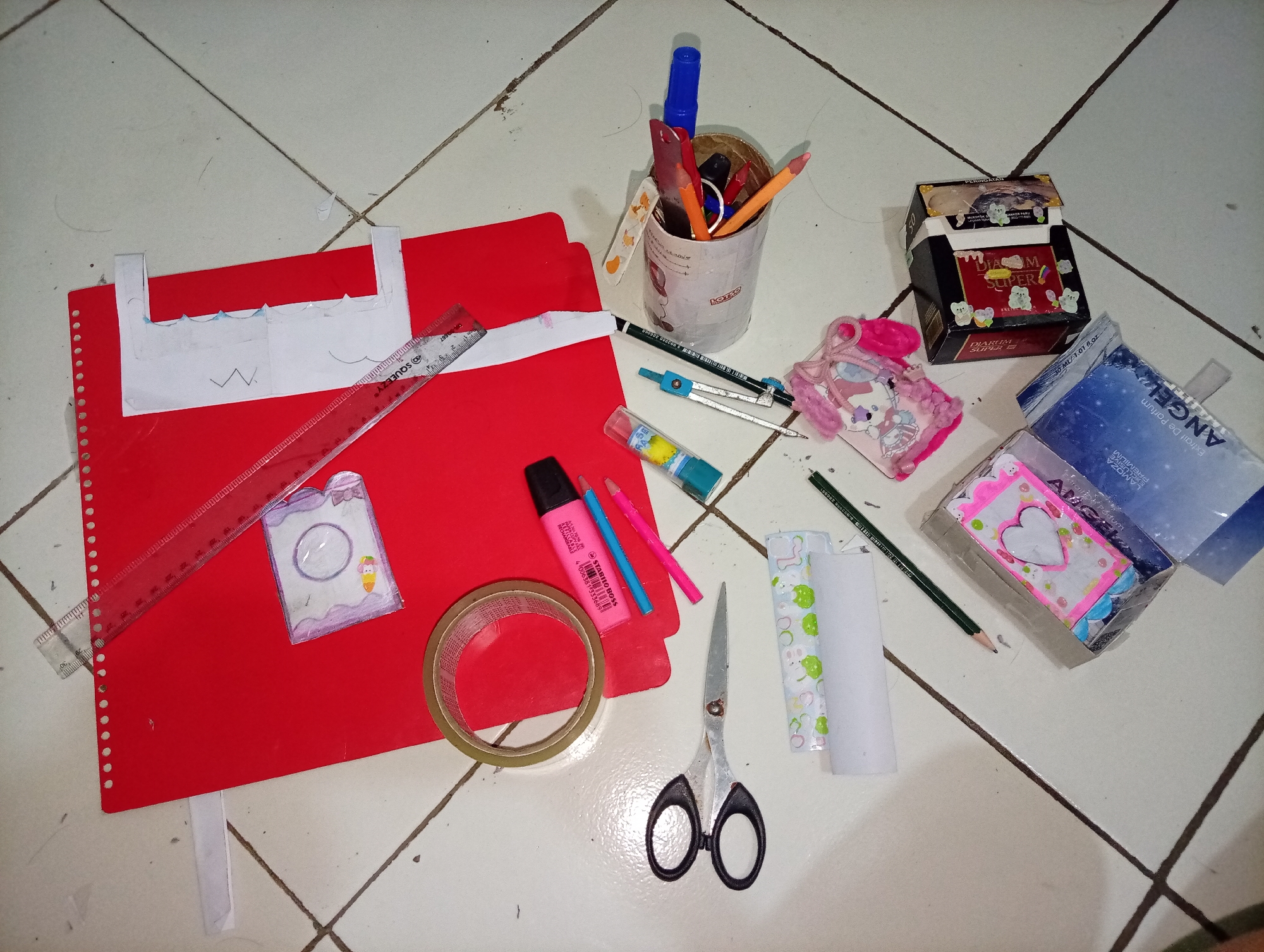 On a tiled floor, likely in a residential setting in Wanajaya, Indonesia, various craft supplies are spread out. A large red folder serves as a base for several items. A ruler, tape measure, roll of tape, scissors, and various writing utensils including markers and pencils are visible. There is also a compass and a glue stick. Several small decorative items, including what appears to be a handmade pouch or card with cartoon characters, are also present. A cigarette pack, decorated with stickers, and an open box, possibly containing more craft supplies or trinkets, are also on the floor. The scene suggests a creative project in progress or recently completed.