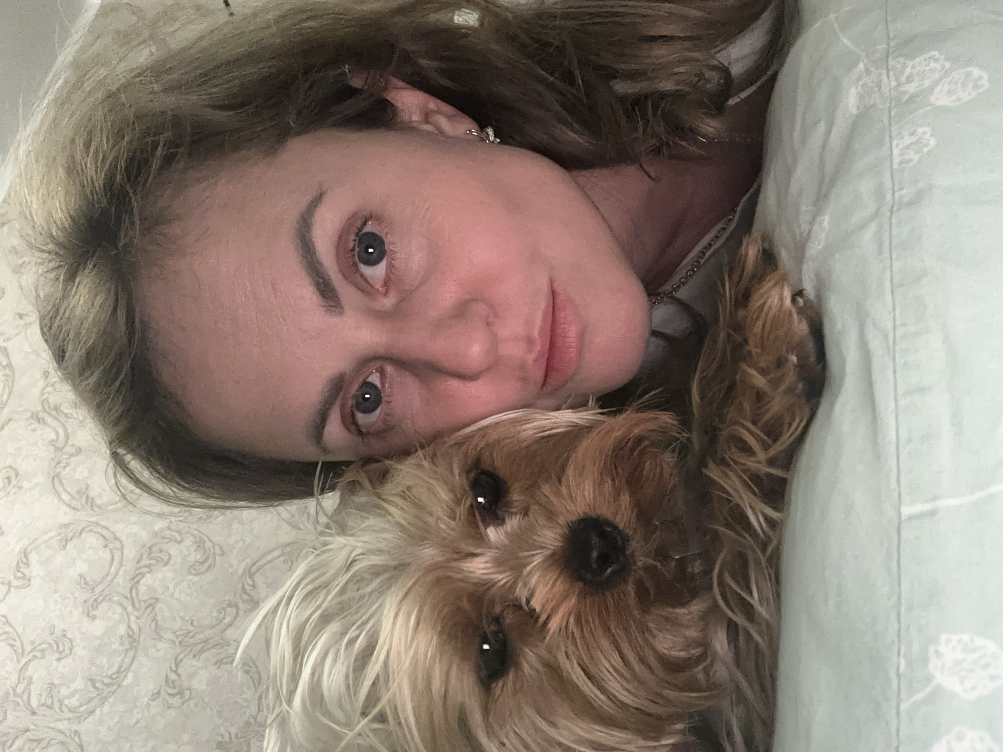 A close-up selfie features a woman lying in bed with her Yorkshire Terrier. The woman is on the right side of the frame, looking towards the camera with a slight smile. Her blonde hair is loose and falls around her face. She has blue eyes and is wearing a small, silver earring. The dog is positioned on the left side of the frame, nestled against the woman's head, with its face prominently displayed. The dog has light brown and tan fur. They are lying on a light green pillow with a white floral pattern. The background is a textured wallpaper with a light grey and white damask pattern. The scene appears to be indoors, likely in a bedroom. The location of Verkhniy Yurt, Russia, is a contextual detail that cannot be visually confirmed from the image alone.