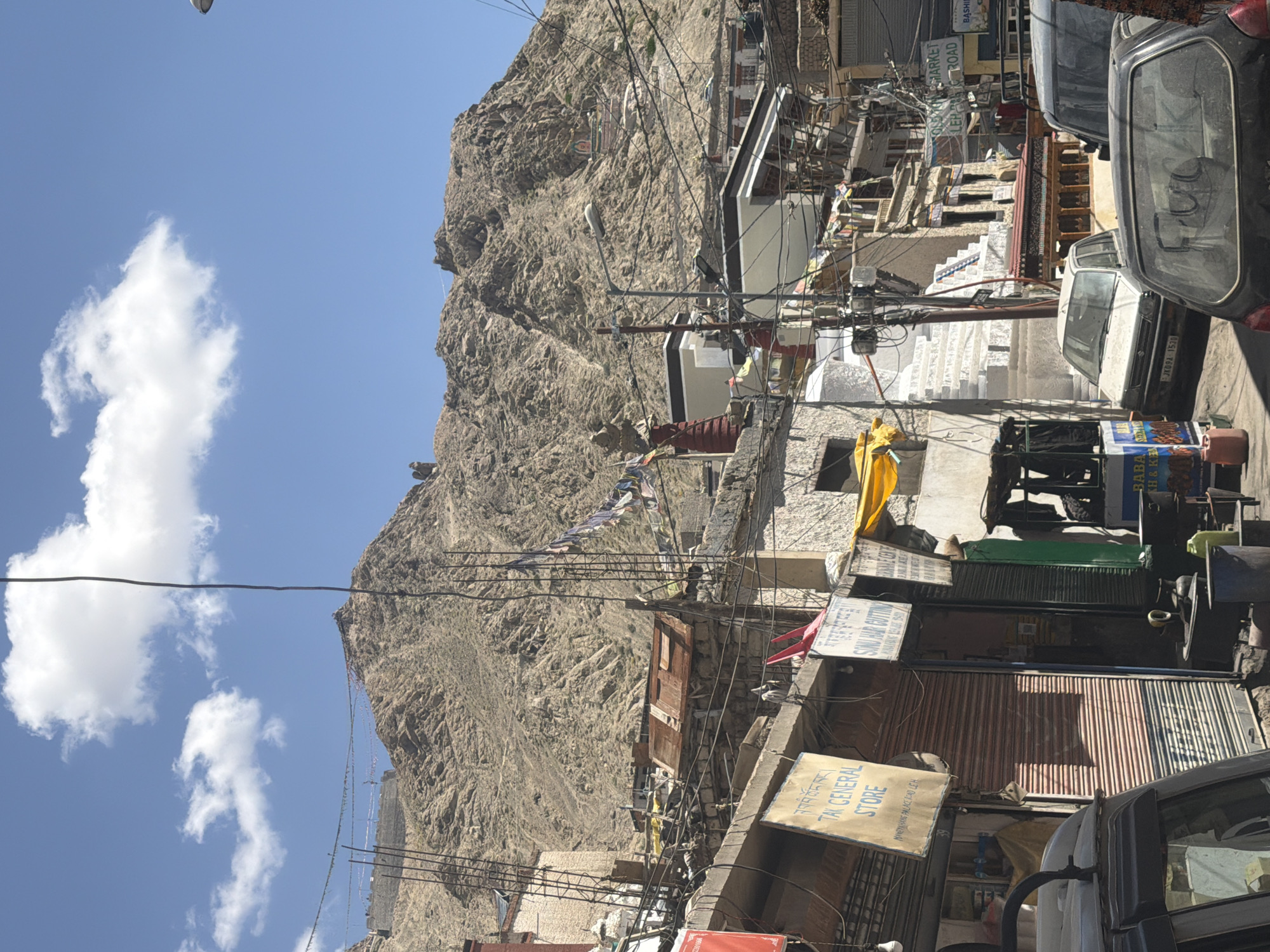 This image captures a street scene in Leh, India, a high-altitude desert city known for its Tibetan Buddhist culture and stunning mountain landscapes. The foreground features a street lined with shops, some with rolled-down shutters, and several vehicles, including a silver car and a black car. Overhead, a complex network of electrical wires crisscrosses the frame, a common sight in many urban areas.
In the background, a majestic, barren mountain rises prominently, its slopes etched with textures and shadows. Prayer flags, a vibrant symbol of Tibetan Buddhism, flutter on lines strung across the scene, adding a splash of color against the muted tones of the mountain and buildings. Structures are visible on the mountain's crest, suggesting a monastery or a fortress. The sky is a clear, bright blue, dotted with fluffy white clouds, indicative of a sunny day.
One of the shops has a sign that reads "TAI GENERAL STORE," with additional text in what appears to be Tibetan script. Another sign indicates "SONU HAIR CUTTING." Further down the street, a sign for "OLD MARKET LEH ROAD" is visible. A white stupa, a Buddhist architectural feature, stands partially visible among the buildings. The overall impression is one of a bustling, yet weathered, urban environment set against a dramatic natural backdrop.