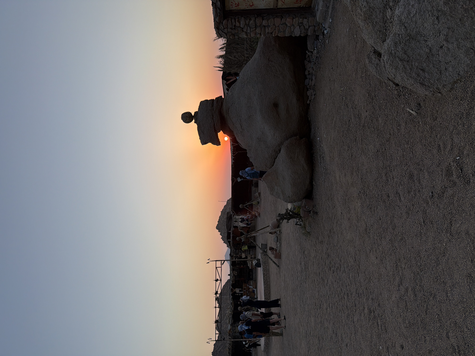 This image depicts a desert scene at sunset, likely in Sharm El Sheikh, Egypt, given the architectural style and arid environment. The foreground is dominated by coarse sand and gravel, with several large rocks, one of which has a unique, bird-like formation topped with a spherical object.
In the mid-ground, a group of tourists, dressed in casual attire and headscarves, are gathered. Some are observing the sunset, while others appear to be engaged in conversation. Behind them, a cluster of rustic structures with thatched roofs and open-air seating areas suggest a tourist attraction or a desert camp. Traditional pottery and wooden tripods are visible, adding to the local ambiance.
The background is characterized by rolling hills and mountains silhouetted against a vibrant sunset sky that transitions from a pale blue at the top to a warm orange and yellow near the horizon. The sun itself is partially obscured by the rock formation, casting a warm glow over the entire scene. The overall atmosphere is peaceful and serene, indicative of a cultural or natural sightseeing experience.