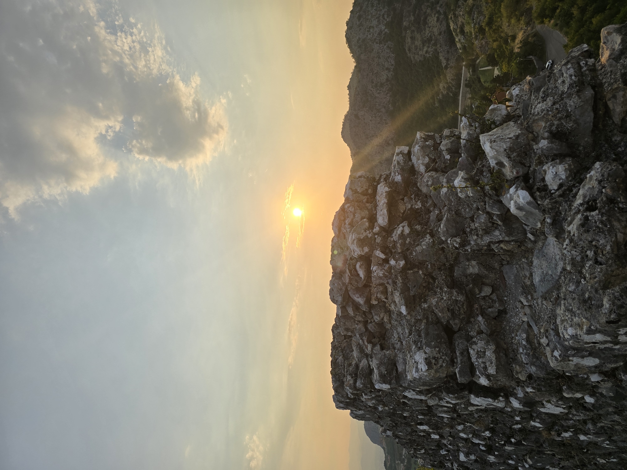 The image shows a close-up of a stone wall, likely part of a historical structure, in the foreground. The stones are rough and irregular, with some appearing mossy or weathered. Beyond the wall, a landscape of rolling hills unfolds under a sky illuminated by a setting or rising sun. The sun, a bright orb, is visible in the upper portion of the frame, casting golden rays across the scene. Clouds are scattered across the sky, tinged with the warm hues of sunset. In the distance, faint outlines of mountains can be seen. A road with a few vehicles and some scattered buildings are visible winding through the valley below. The overall impression is one of a serene, natural landscape at dusk or dawn, with the ancient stone structure adding a sense of history and timelessness. The location is identified as the city of Medun, Montenegro.