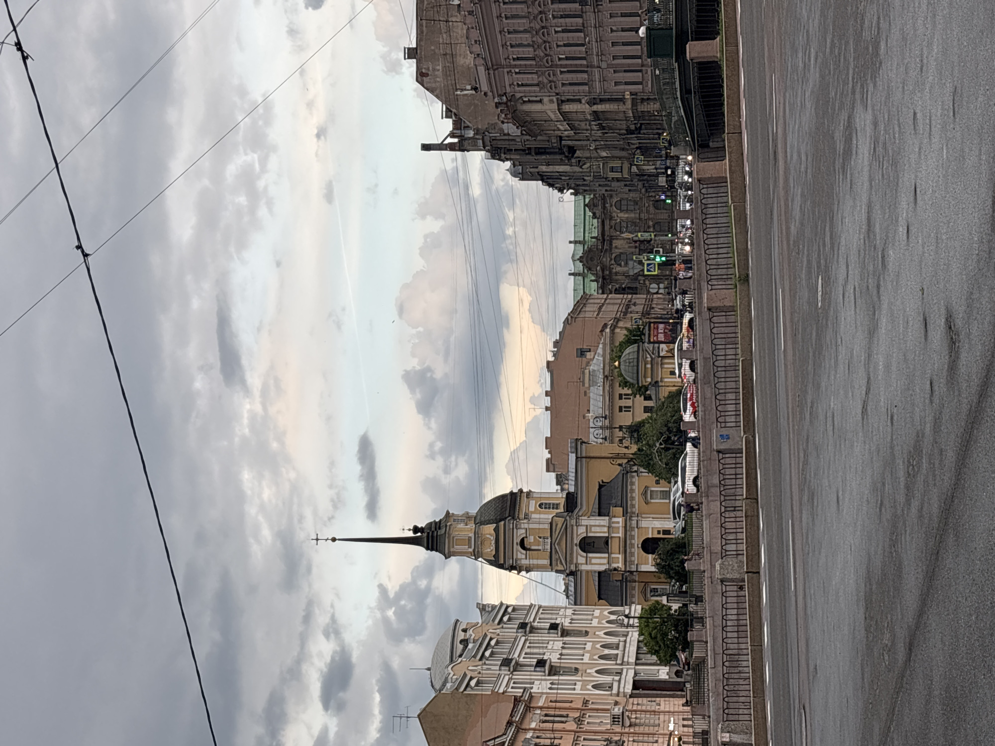 This image captures an urban scene in Saint Petersburg, Russia, specifically focusing on the area around the **Church of the Holy Great Martyr and Healer Panteleimon** (Пантелеймоновская церковь), located near the Fontanka River.
In the center of the frame, the **Church of the Holy Great Martyr and Healer Panteleimon** stands prominently, distinguished by its pale yellow facade, a tall, slender black spire topped with a golden orb and a cross, and a dome situated below the spire. To the left of the church, a multi-story, light-colored historic building with numerous windows and decorative elements lines what appears to be an embankment or street. Further to the right and in the background, a collection of classical-style buildings with varying shades of grey and brown, some featuring green roofs, completes the historic cityscape.
The immediate foreground consists of a wide expanse of dark, wet asphalt, likely a street or public square, marked with white traffic lines. A low stone parapet with an ornate black railing separates the road from the background structures and what is presumed to be the Fontanka River. Behind this barrier, several vehicles, including cars of various colors (e.g., a white SUV), are visible, some appearing parked while others may be in motion. Faintly discernible pedestrians can be seen along the sidewalks and near crosswalks. On the far right, a dark green ornate bridge structure, consistent with the **Panteleimonovsky Bridge**, crosses an unseen waterway. Traffic lights and streetlights are visible at various points, indicating an active urban environment.
The sky is heavily overcast with numerous shades of grey and white clouds, suggesting a cloudy day with soft, diffused light. Multiple dark overhead utility or tram wires crisscross the upper portion of the sky. The overall impression is a detailed snapshot of a historic European city on a day with subdued lighting.