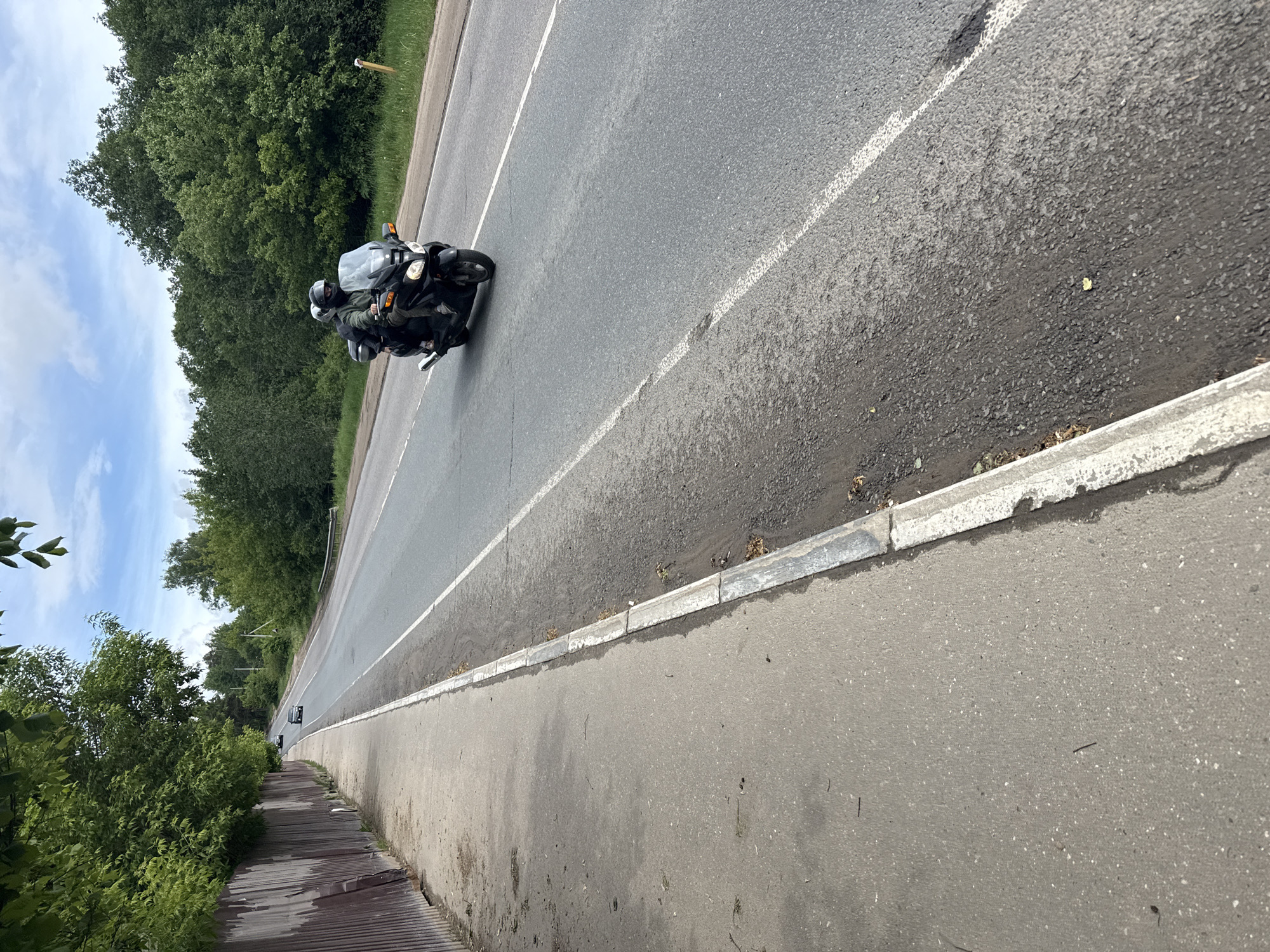 The image shows a two-person motorcycle traveling along a road in Dmitrov, Russia. The road has white lane markings and a curb separating it from a sidewalk. There are trees lining the road on both sides. The sky is partly cloudy. A vehicle is further down the road behind the motorcycle. The view appears to be taken from a low angle, and the image is rotated.