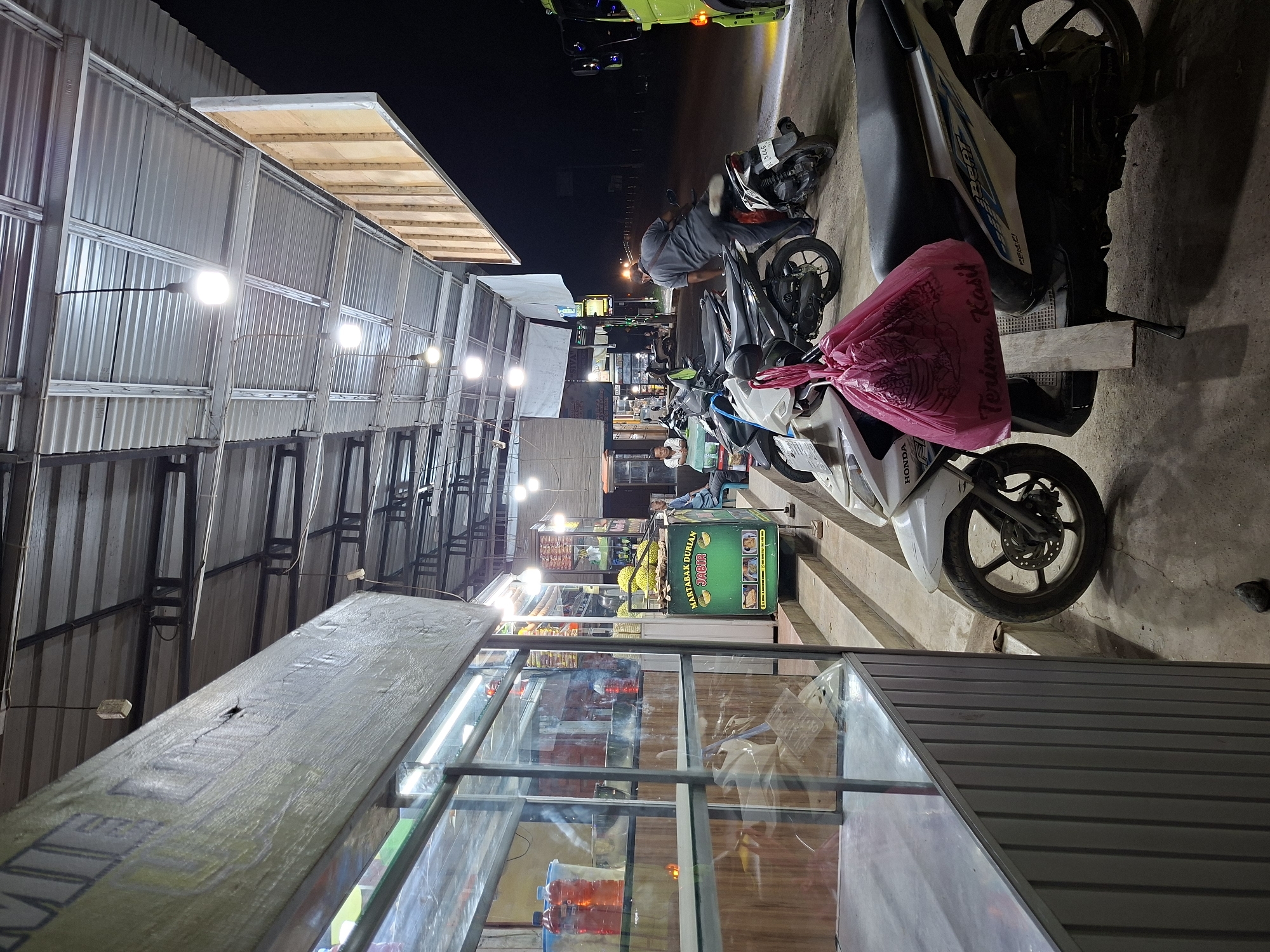 The image captures a nighttime street scene in Keudegeudong, Indonesia, likely a bustling food vendor area. The setting is illuminated by numerous bare light bulbs strung along makeshift structures, casting bright light and shadows.
On the left side, a series of stalls with corrugated metal roofs and walls line what appears to be a walkway or side of a street. These structures are basic, utilizing metal framing.
In the foreground and central part of the image, several motorcycles, mostly scooters like a Honda Beat, are parked. One white scooter has a pink plastic bag tied to its seat, featuring the text "Terima Kasih" (Thank You) in white script. Another scooter's license plate shows part of "5875 BQ".
Towards the middle ground, three individuals are visible. One man in a grey shirt is leaning over a parked motorcycle. Another person is seated behind a vibrant green food cart. This cart prominently displays the text "MARTABAK DURIAN JADUL" and shows several durian fruits, indicating it sells durian-based pancakes or sweets. Behind this cart, another vendor stand with glass displays is visible, stocked with various items. Further down the street, streetlights and additional vehicles can be seen, suggesting a continuous road.
The overall scene depicts a typical evening atmosphere at an outdoor market or food stall area, characterized by vendors operating under artificial lighting for late-night customers.