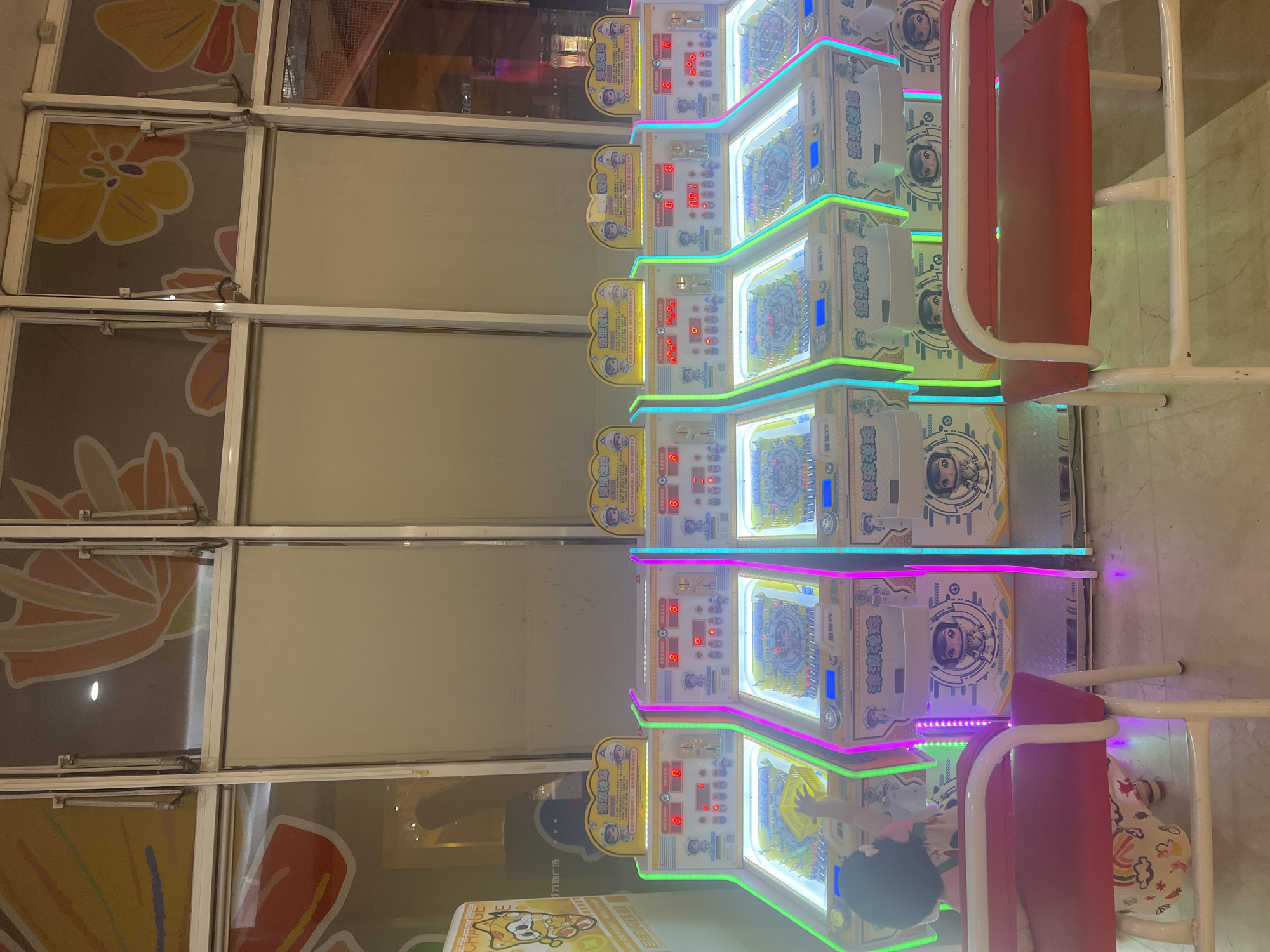 In Liandu District, China, a young child, viewed from the back with dark hair and wearing a patterned outfit, is playing a coin-operated game. The child's left arm is raised, reaching towards the illuminated playing field of a brightly lit arcade machine. The machine is one of a row of five identical units, each featuring colorful LED lighting along its sides in green, pink, and blue. The machines are adorned with cartoon characters and Chinese text. Two red benches with white metal frames are positioned in front of the machines, one partially obscured by the child. The floor is tiled in a muted tone, and the background features large windows with decorative floral patterns.