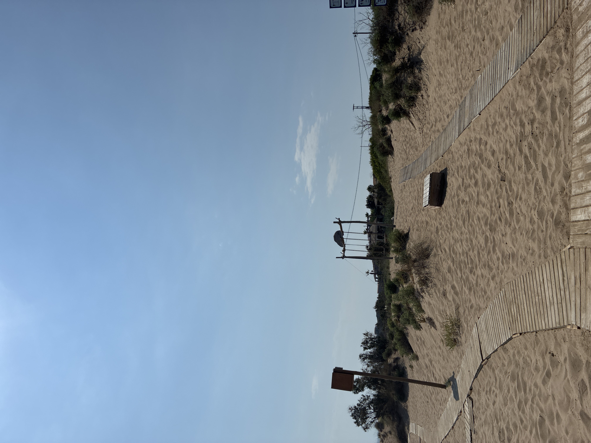 This is a photograph of a sandy beach with wooden boardwalks. The boardwalks are laid out in a curved path across the sand, leading up a gentle dune. There is some sparse vegetation growing on the dunes, including bushes and a few trees. In the distance, there are buildings and a swing set. The sky is blue with a few scattered clouds. The image appears to have been taken on a sunny day. The location is likely a beach resort or park area in Zakynthos Municipal Unit, Greece, given the typical landscape of the region and the presence of amenities like boardwalks and a swing set. There are no people visible in the image.