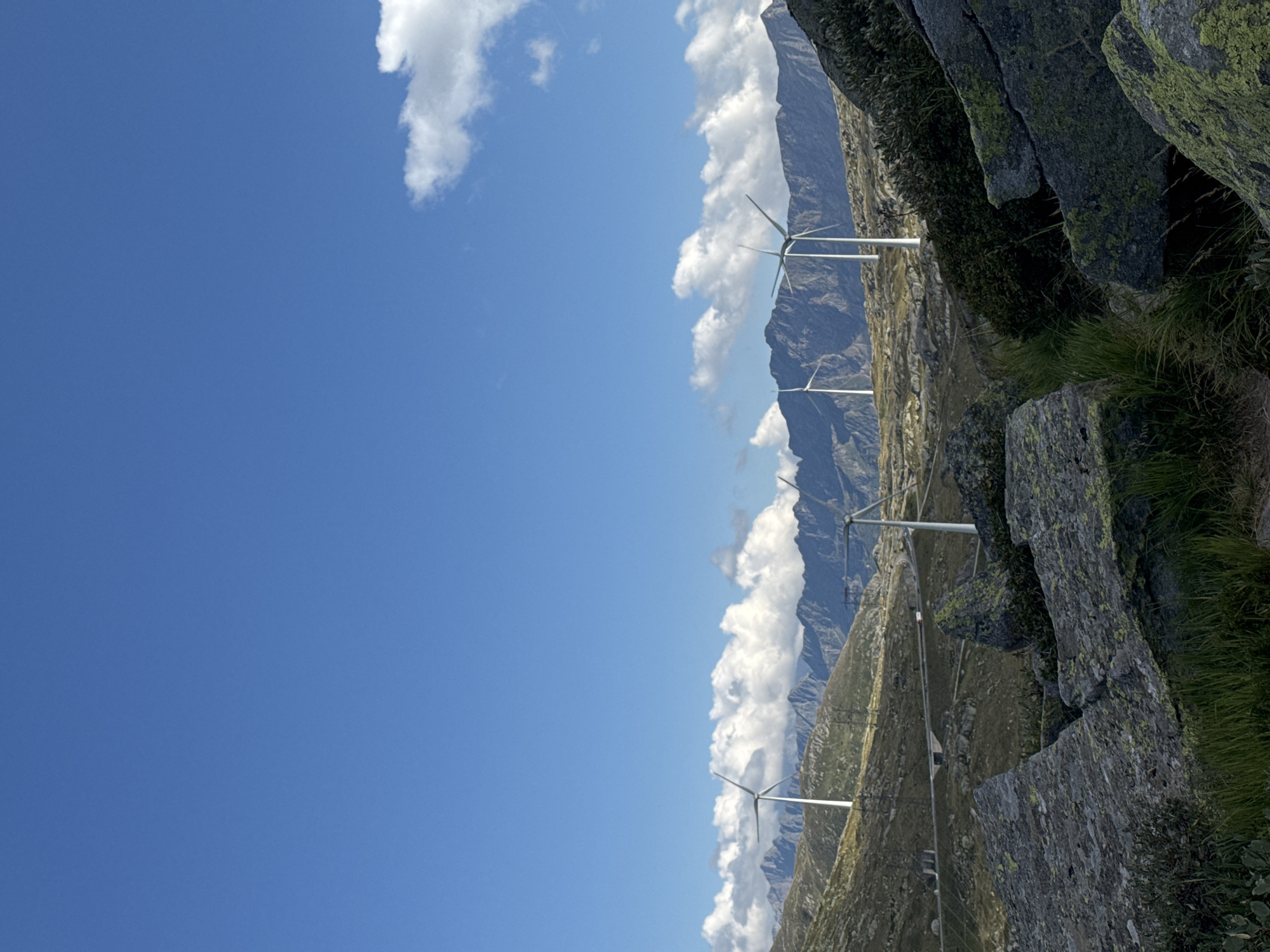 The image depicts a mountainous landscape with several wind turbines visible on a ridge under a bright blue sky with scattered white clouds. In the foreground, large moss-covered rocks and patches of dry grass dominate the scene. A winding road with a tunnel can be seen further in the distance, traversing the hilly terrain. The environment appears rugged and natural, consistent with a high-altitude location. The specific location is the Gotthard Pass near Airolo, Switzerland, an area known for its wind farm. There are no people or animals visible in the image.