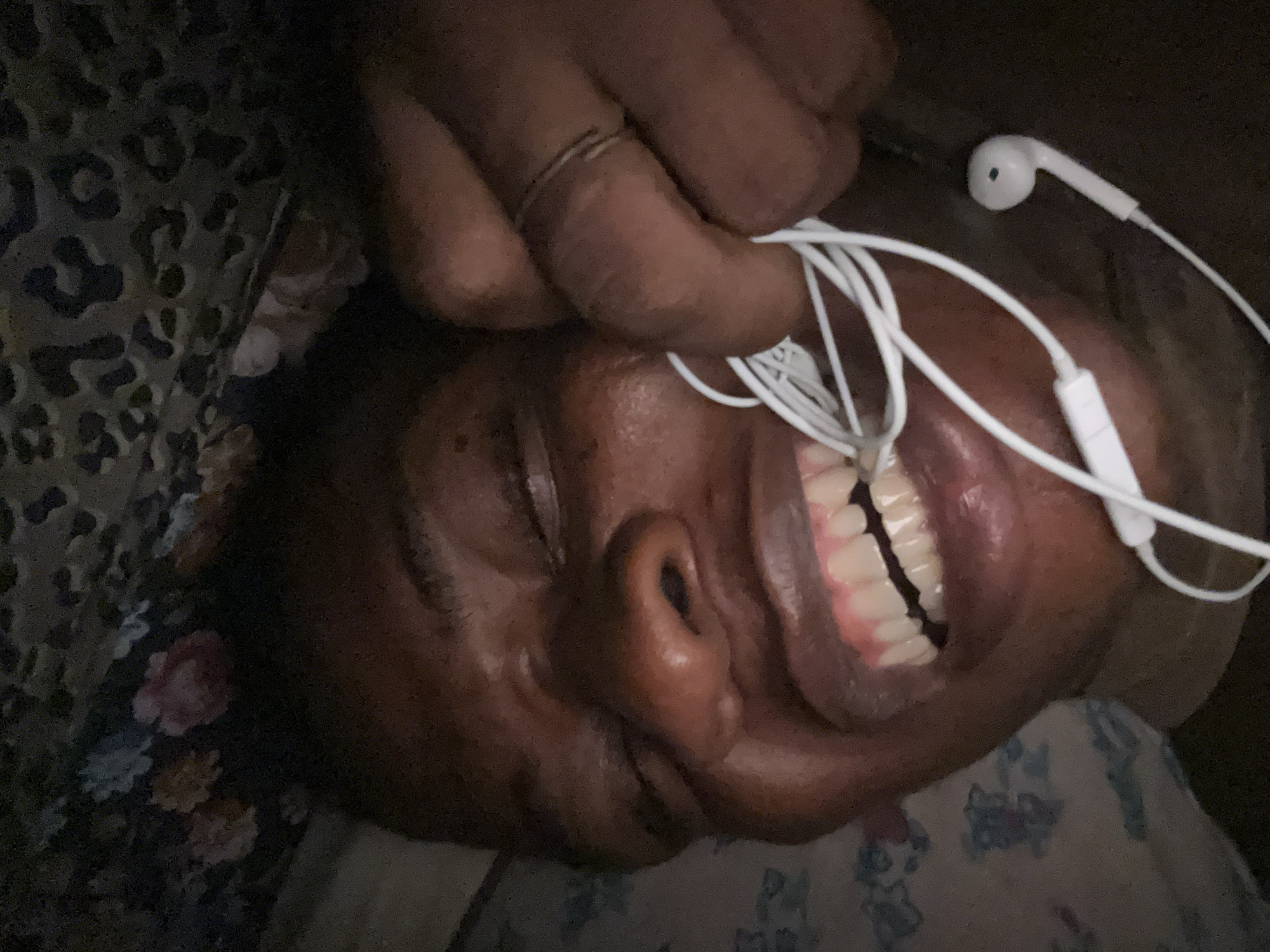 The image shows a person with dark skin lying down, possibly on a bed, with white earphones tangled around their face. Their eyes are closed, and their mouth is open, revealing teeth. A hand, also with dark skin, is holding the earphones. The background includes what appears to be a floral patterned pillowcase and possibly a patterned garment. The location is unknown.