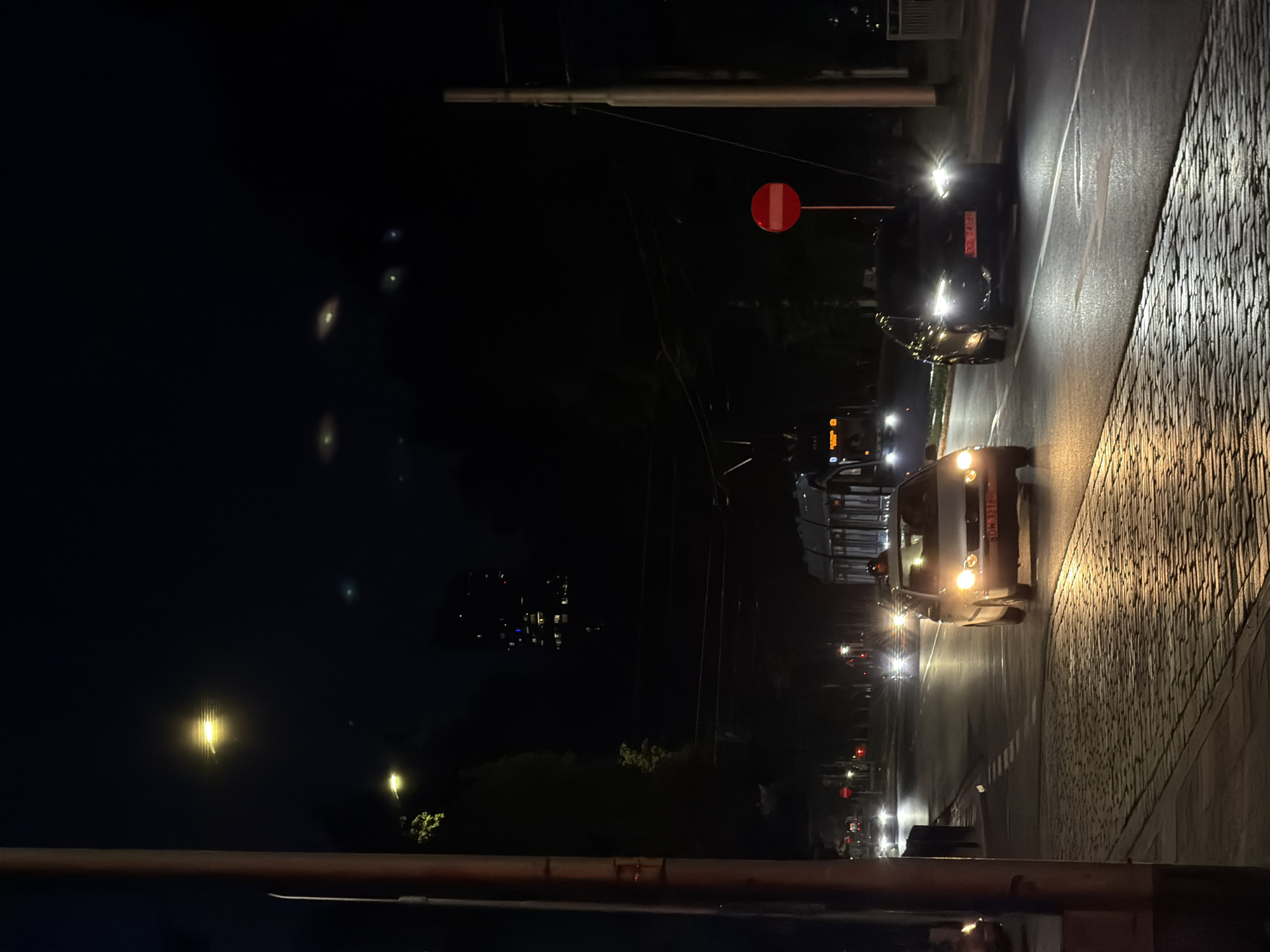 An evening street scene in an urban environment is depicted. The foreground features a cobblestone sidewalk adjacent to a paved road. Several vehicles are visible on the road, some with their headlights on, illuminating the street and the wet asphalt. In the background, a tram or light rail vehicle is present, along with streetlights and distant building lights. A red circular sign with a horizontal white bar is mounted on a pole along the roadside. The overall impression is a dimly lit, nighttime city street with active traffic.