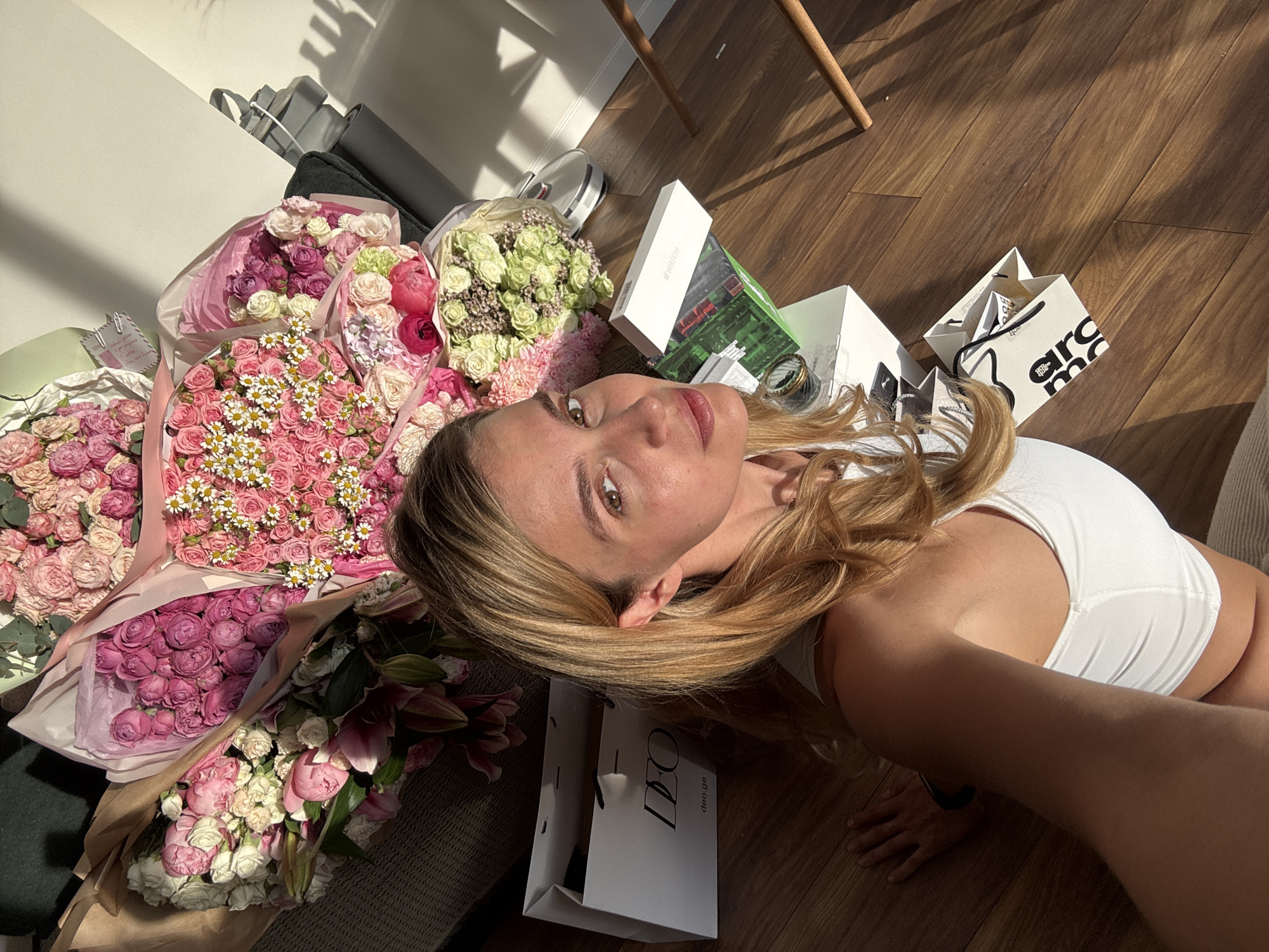 A woman with blonde hair, wearing a white top, is lying on a wooden floor, surrounded by several bouquets of flowers in shades of pink, purple, and green. There are also several shopping bags and gift boxes scattered around her. The scene is lit by sunlight. The location is Unknown, Unknown.