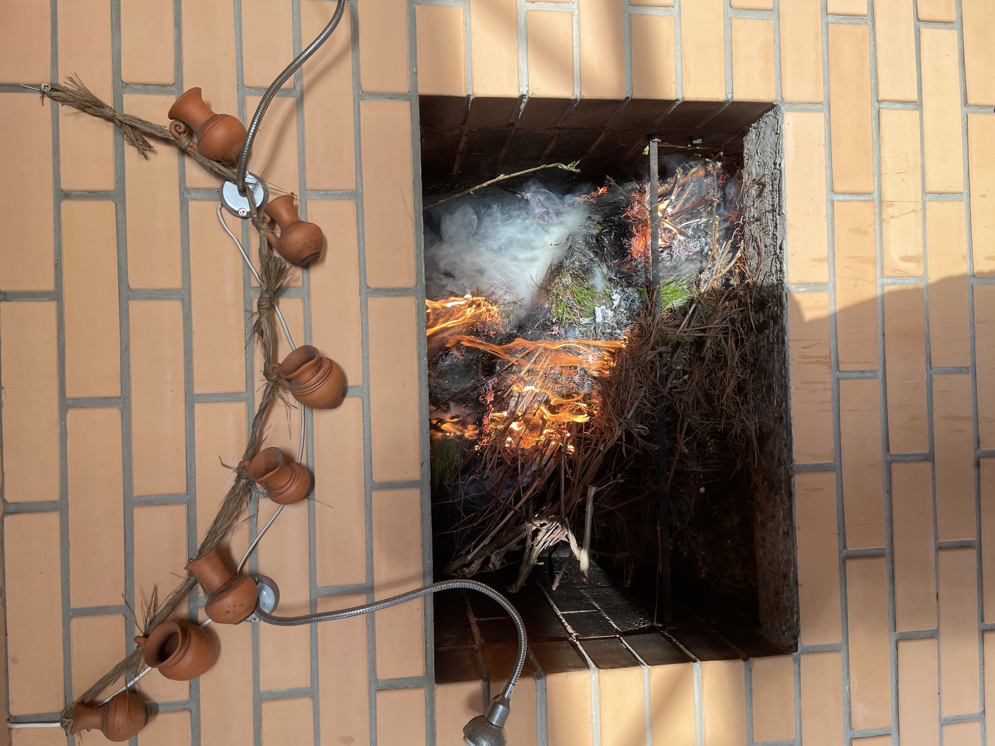 The image shows a brick structure, likely a fireplace or oven, with a fire burning inside. The structure is made of light brown bricks laid in a running bond pattern. Inside the opening, flames are visible, along with smoke and what appears to be burning brush and small branches. To the left of the structure, decorative clay pots are attached to a twig-like vine, which is itself attached to the brick wall with a silver, flexible lamp. The location is Лесной Пейзаж Кп (шарапово, Russia.