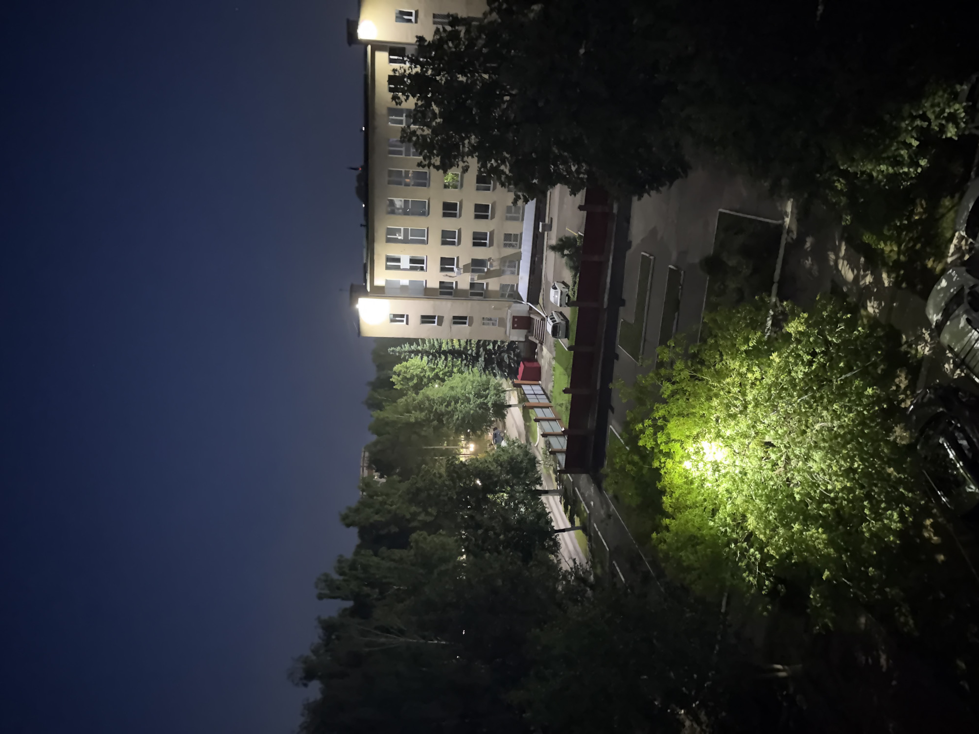 An eye-level, full outdoor shot captures a building complex at night in an urban setting. The sky is a deep, dark blue, suggesting twilight or early night. A multi-story building with numerous windows and brightly lit sections is the focal point. The building is beige or light-colored with a flat roof topped with lights. In front of the building, there is a paved area with two cars, a white sedan and a light-colored SUV, parked. A reddish-brown fence encloses a grassy area to the left of the building, where a red shed is visible. Trees of various sizes and densities surround the building and extend into the background, some illuminated by artificial lights. In the foreground, a dense cluster of green trees is brightly illuminated by a spotlight, casting a strong green glow. The ground in the foreground and middle ground appears to be a mix of paved surfaces and grass. Several vehicles are visible in the foreground, mostly obscured by darkness and foliage.