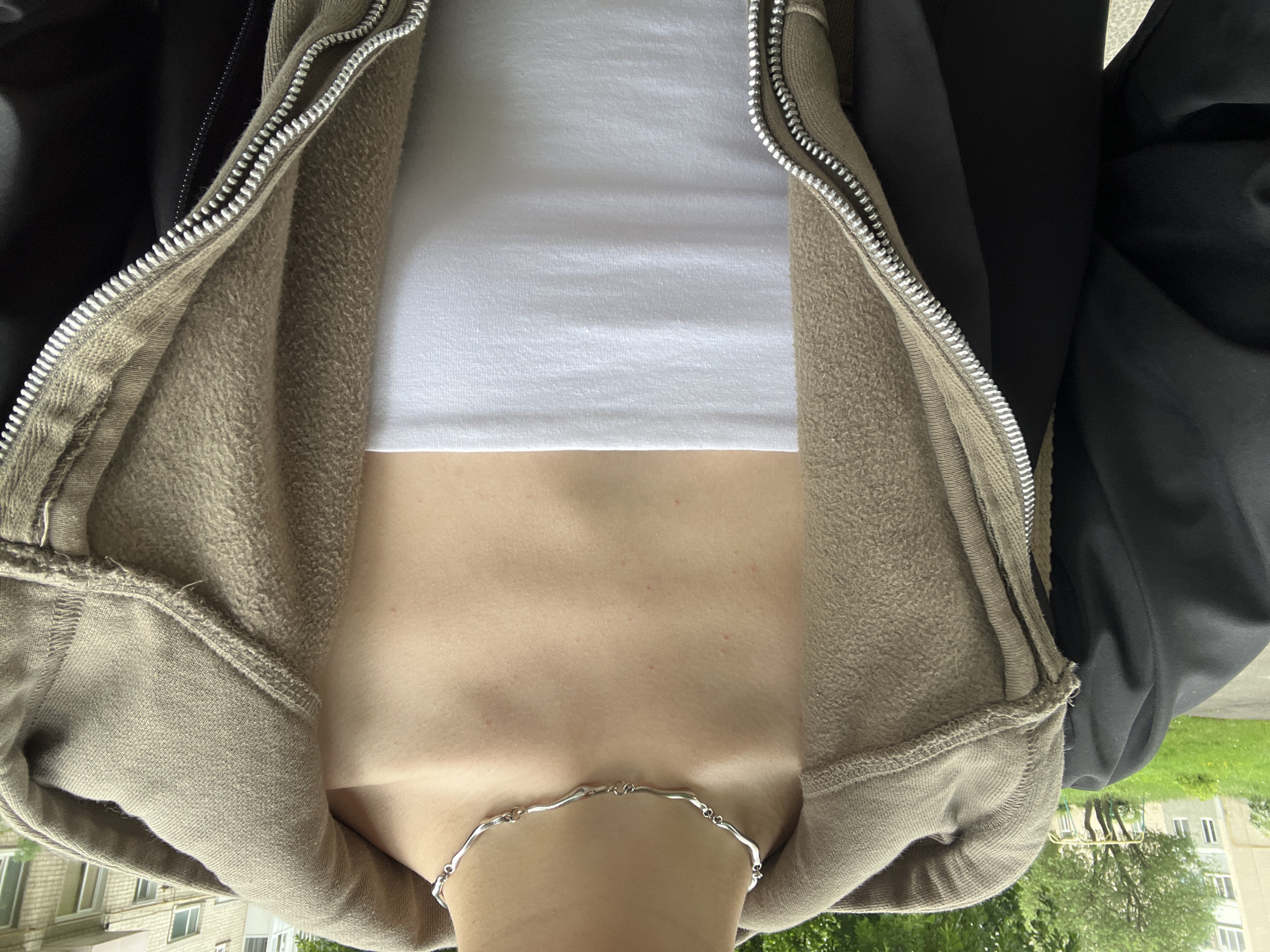 A close-up, slightly tilted image shows a person from the chest up. They are wearing a white, cropped tank top under a partially open, tan-colored hoodie with a silver zipper. Over the hoodie is a black jacket or coat. A silver necklace rests around the person's neck. In the blurred background, there is greenery and the side of a building with windows. Based on the metadata, the location is Ternopil, Ukraine.