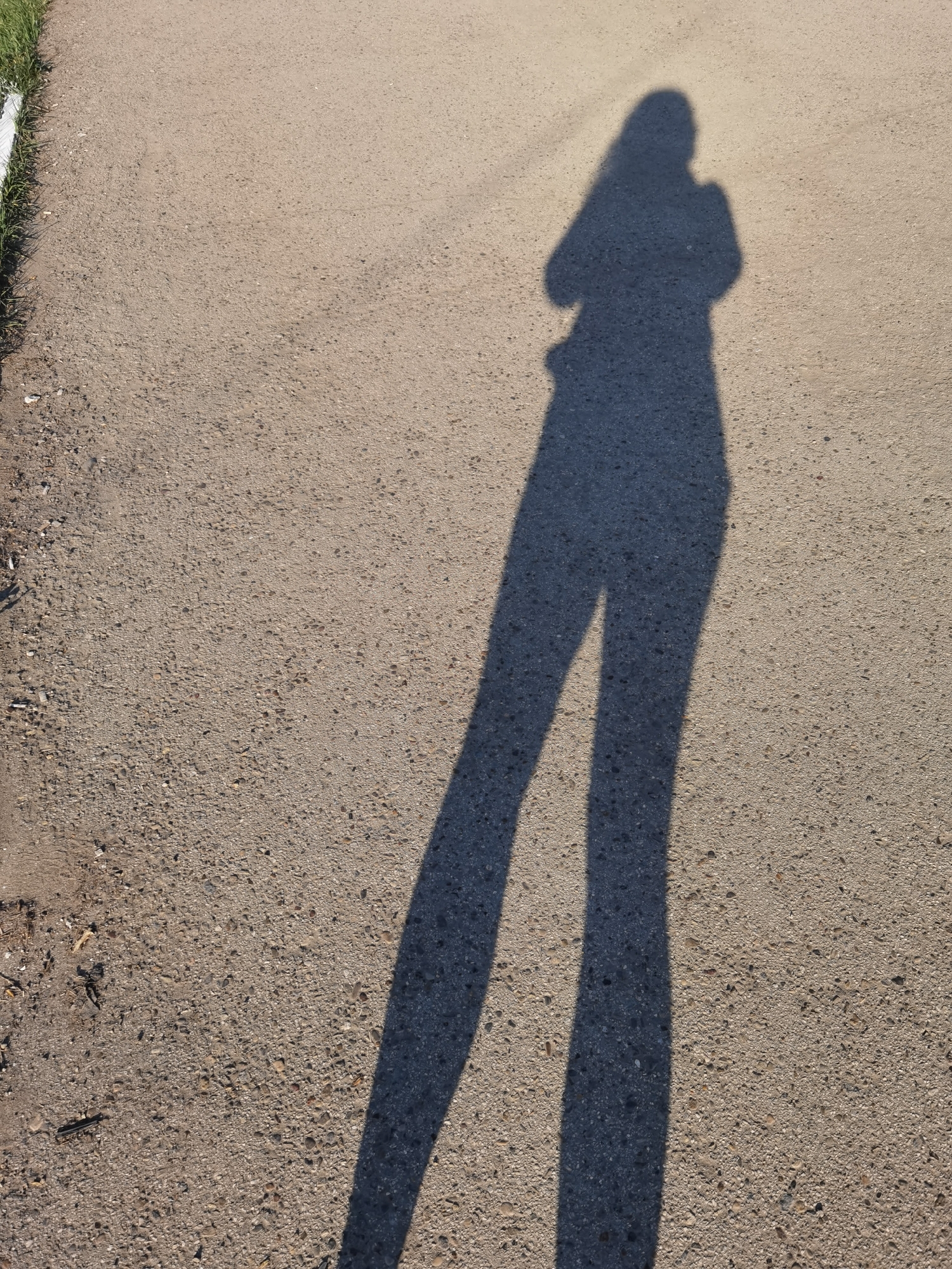 The image depicts a shadow of a person on an asphalt surface. The shadow is elongated, suggesting a low sun angle. The person's silhouette indicates they are likely standing upright, with arms crossed or holding something in front of them. The asphalt surface appears to be part of a road or walkway. The left edge of the image shows a white curb and some green grass. The location is unknown.