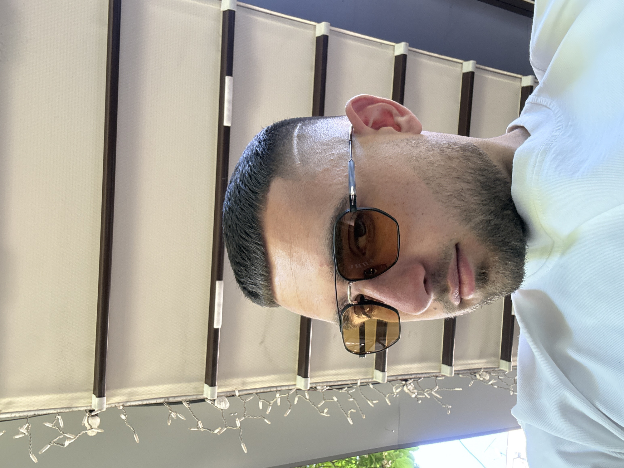 The image shows a man wearing sunglasses and a white shirt. He is positioned in front of a structure with dark brown vertical beams and white panels, likely a canopy or awning. There are decorative lights hanging beneath the structure. The location is potentially Stara Zagora, Bulgaria, but there are no clear indications of this within the image.
