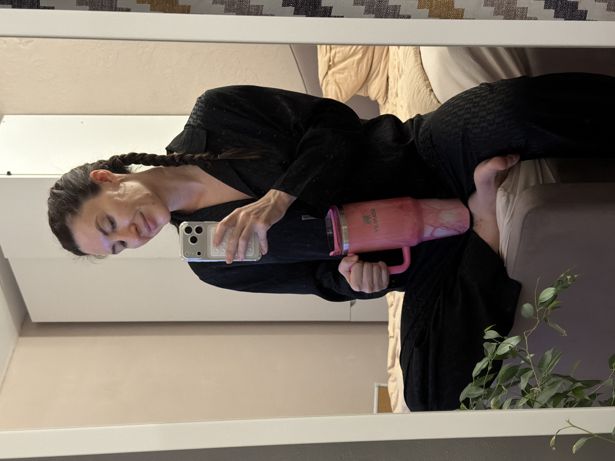 A single adult female, estimated to be in her 20s to 30s, is depicted in a mirror selfie taken indoors. She has dark hair styled in two braids and is wearing black long-sleeved pajamas with a subtle embossed pattern. She is seated, with her bare right foot visible, and holds a silver-backed smartphone with a clear case in her right hand, positioned to capture her reflection. Her left hand holds a large, pink Stanley tumbler with a handle, exhibiting a textured or marbled finish. She gazes downwards towards the phone with a faint smile.
The immediate background, reflected in the mirror, includes a light beige wall, a white wardrobe or cabinet, and a section of a bed with light beige bedding. A green leafy plant is visible in the lower left foreground, partially obscured by the mirror frame. On the far right, a segment of a patterned rug or carpet with geometric designs in brown, white, and purple hues is discernible. The environment suggests a domestic setting. The specific location within Perm, Russia, cannot be determined from the visual information presented in the image.