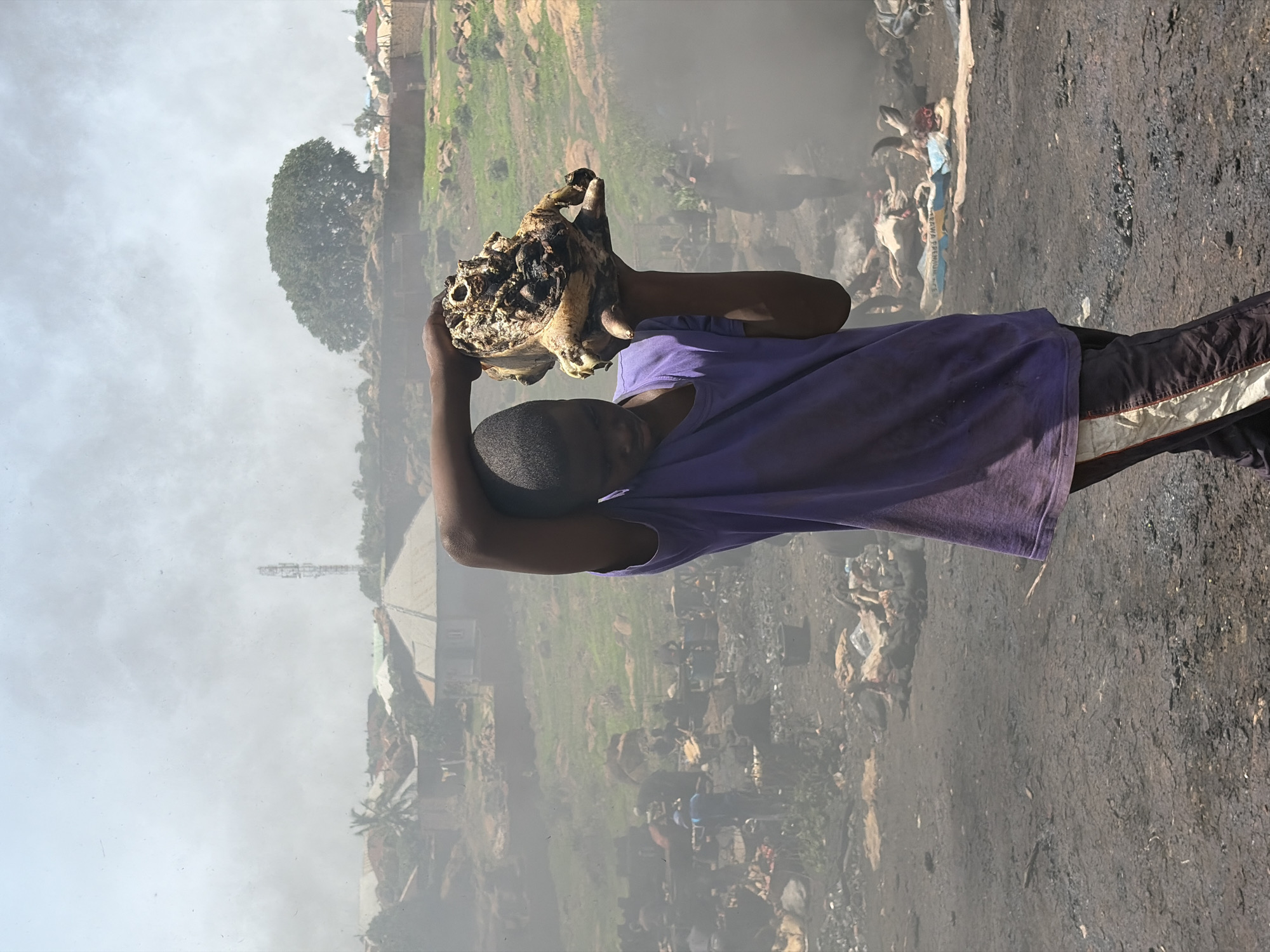A person wearing a purple shirt is carrying what appears to be a charred animal skull above their head with both hands. The location appears to be an outdoor area in Jos, Nigeria. The environment is hazy, potentially due to smoke. In the background, there are structures and greenery suggestive of a residential or urban area. In the foreground, there's a dirt or gravel ground surface. There are also pieces of trash and debris visible in the background and foreground.