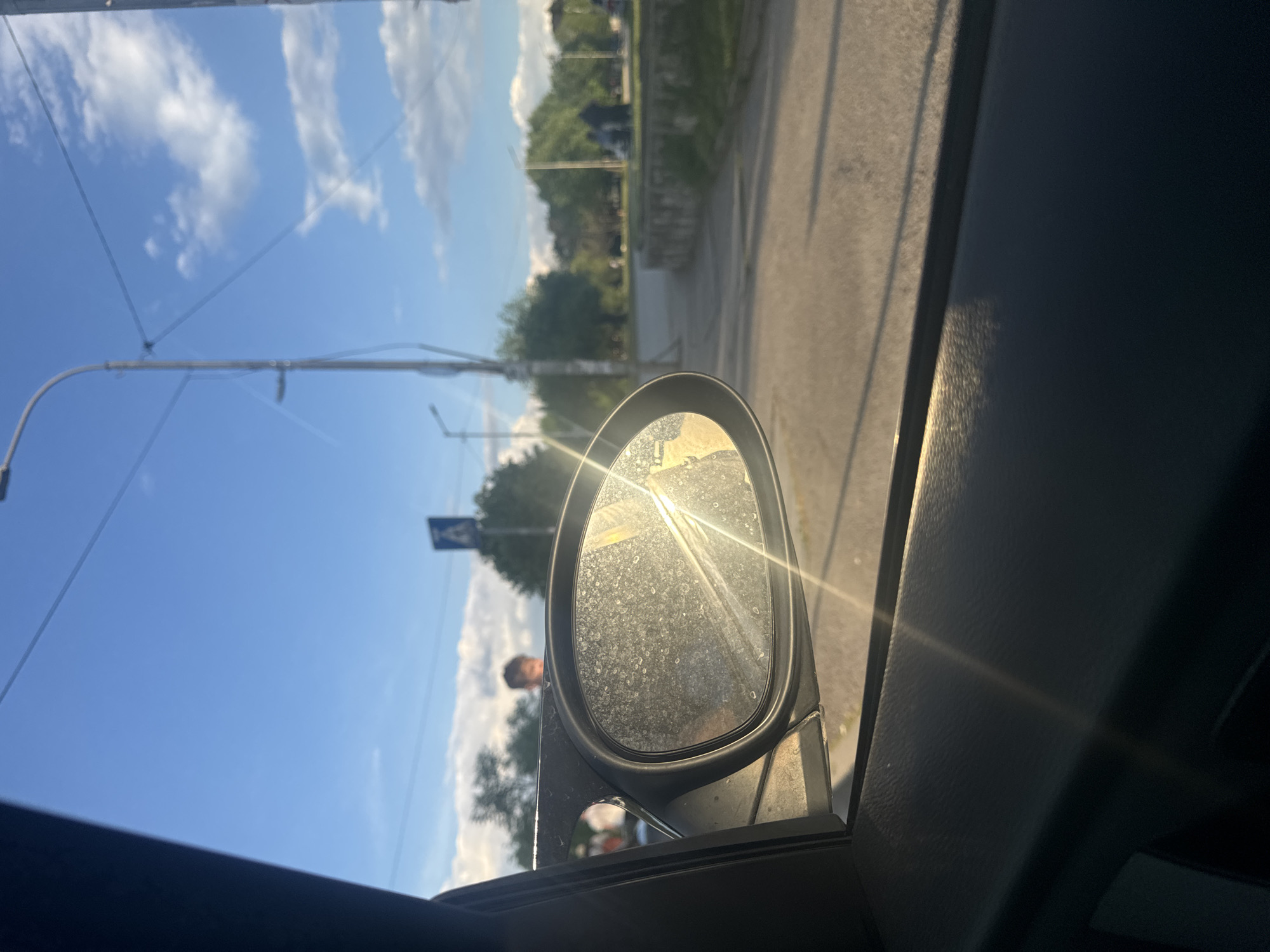 The image shows a side mirror view from inside a car, likely in Sofia, Bulgaria. The side mirror is oval-shaped and displays a bright glare from the sun. Dust and small particles are visible on the mirror's surface. The outside view through the mirror includes a blue sky with clouds, a street lamp, trees, a road, and a pedestrian crossing sign. A blurry figure of a person is reflected in the mirror. The interior of the car, including the door panel, is visible. The day appears to be sunny.