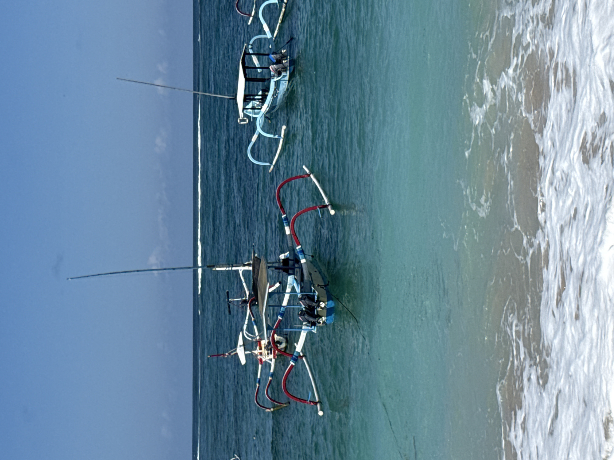 This image depicts several Balinese jukung boats anchored in the turquoise waters off the coast of Kuta Selatan, Indonesia. The boats, characterized by their outriggers and brightly colored decorations, are a common sight in this region. The water is clear, with gentle waves lapping at the shore in the foreground. The sky is a vibrant blue with a few scattered clouds. The scene suggests a calm day at sea, with the boats awaiting their next voyage.