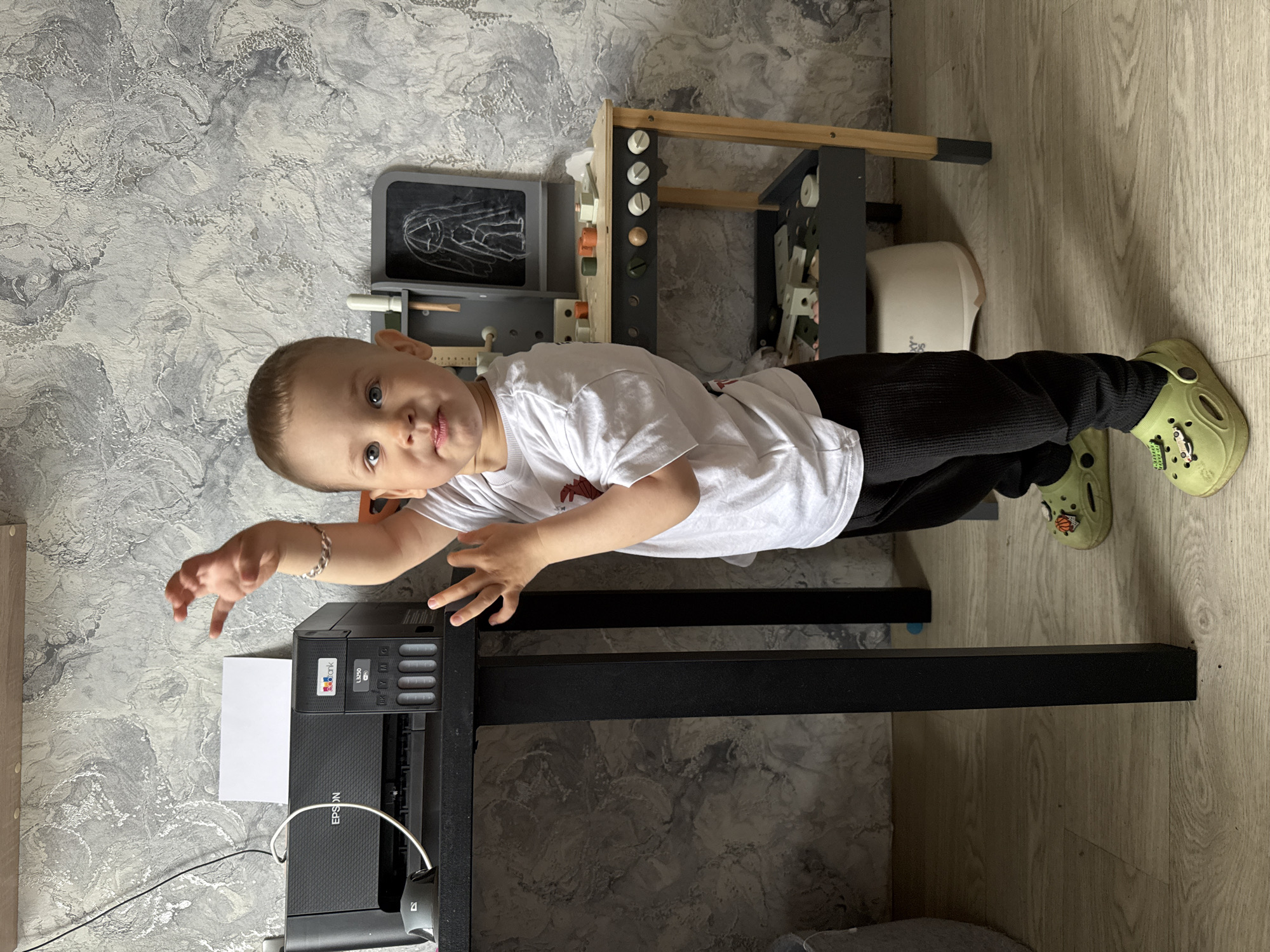 A young child, estimated to be between 1 and 3 years old, is positioned in front of a black printer, likely an Epson model, placed on a table with a black frame. The child wears a white t-shirt and black pants, and is shod in light green Crocs. The location appears to be indoors, evidenced by the marble-patterned wallpaper and wood-toned floor. To the right of the child are wooden toys. The image's orientation is rotated 90 degrees counter-clockwise. The city location, Rusanovo, Russia, is derived from external information, not visually evident in the image.