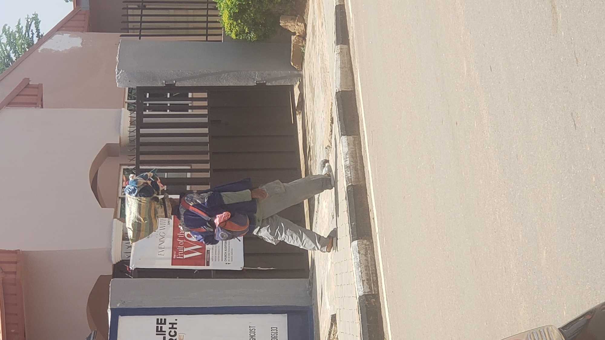 In Narayi, Nigeria, a person walks along a sidewalk carrying a large, bundled load balanced on their head. The individual is wearing loose-fitting grey trousers and a dark blue jacket over a lighter colored long-sleeved shirt. A backpack with red and blue elements is worn on their back. They are walking from left to right across the frame. Behind the person, a dark metal gate stands in front of a building with beige walls and a brown roof. A sign is visible to the left of the gate, featuring text including "LIFE," "RCH," and "HIGHCOST." To the right of the gate, a green bush and some rocks are present. The sidewalk is paved with interlocking bricks, and a road with a black and white curb runs in the foreground. The sky is visible at the top of the image, appearing overcast.