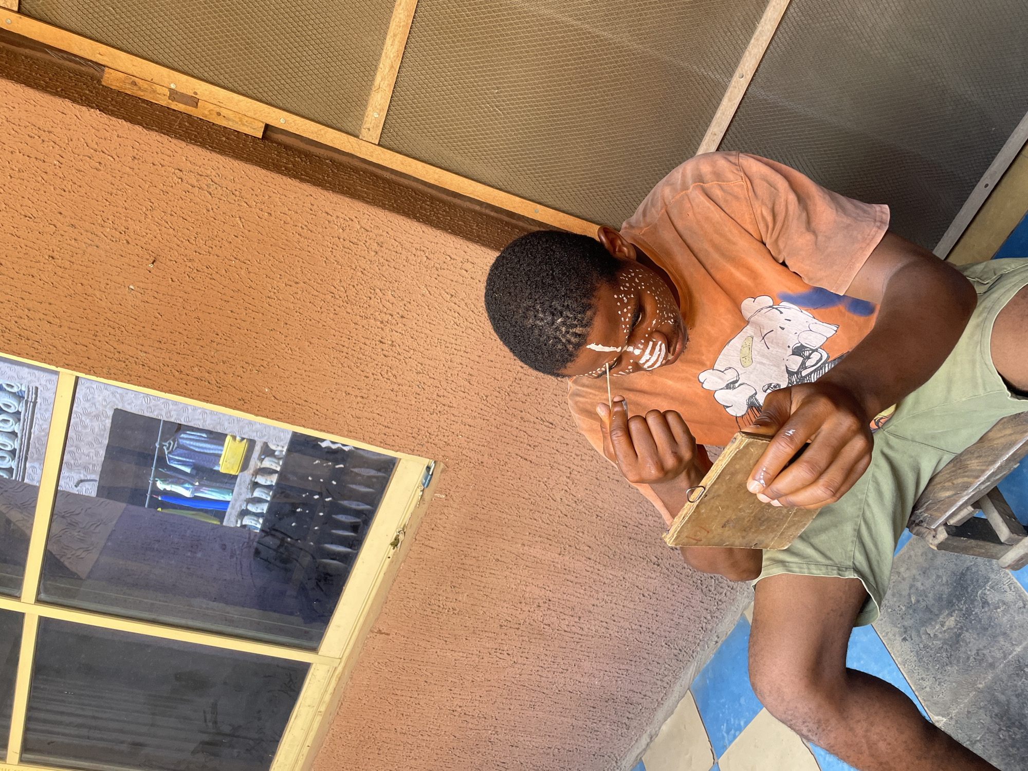 A young Black man with white paint on his face is seated in a room with a textured orange wall. He wears an orange t-shirt and green shorts. He is holding a small, rectangular wooden object in his left hand and a thin stick in his right hand, as if he is working on a project. A window with a yellow frame is visible in the wall, reflecting clothes hanging on a rack. Overhead, there is a screen in a wooden frame. The floor has a blue and grey tiled pattern. The scene appears to be set indoors, possibly in a residence in Nkpor, Nigeria, though there is no definitive evidence of the city present in the image.