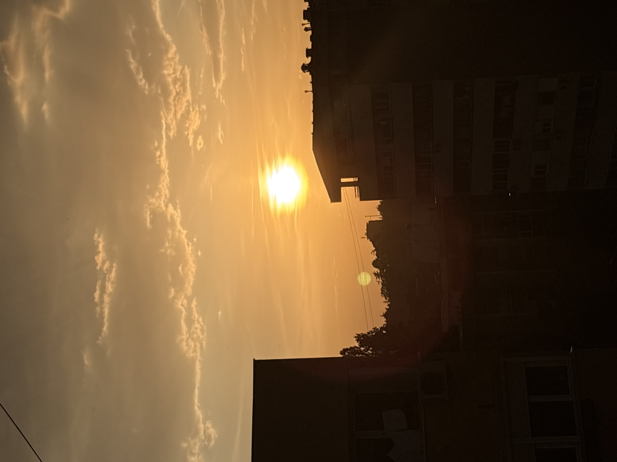 The image shows a sunset in Belgrade, Serbia. The sun is low in the sky, appearing as a bright orb partially obscured by the silhouette of a tall building. Clouds are visible in the sky, rendered in shades of orange and yellow due to the sunset. In the foreground, parts of other buildings are also visible, appearing darker and more silhouetted. Trees can be seen near the buildings. The perspective is from a slightly low angle, looking upwards towards the sun. The scene is urban, dominated by buildings and the setting sun.