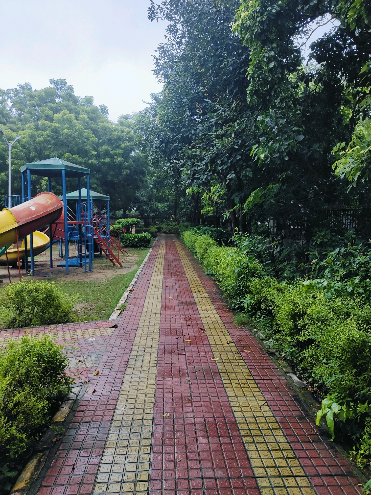 The image depicts a pedestrian walkway made of red and yellow tiles, leading into a park with lush greenery. On the left, there is a children's play area with a yellow and red slide, a blue and green structure with stairs, and a small green gazebo. The path is lined with manicured bushes on both sides, and tall trees form a canopy overhead. The sky is overcast, suggesting a cloudy day. The scene is devoid of people. This park is located in Gurgaon, India, a bustling city known for its modern infrastructure and green spaces.