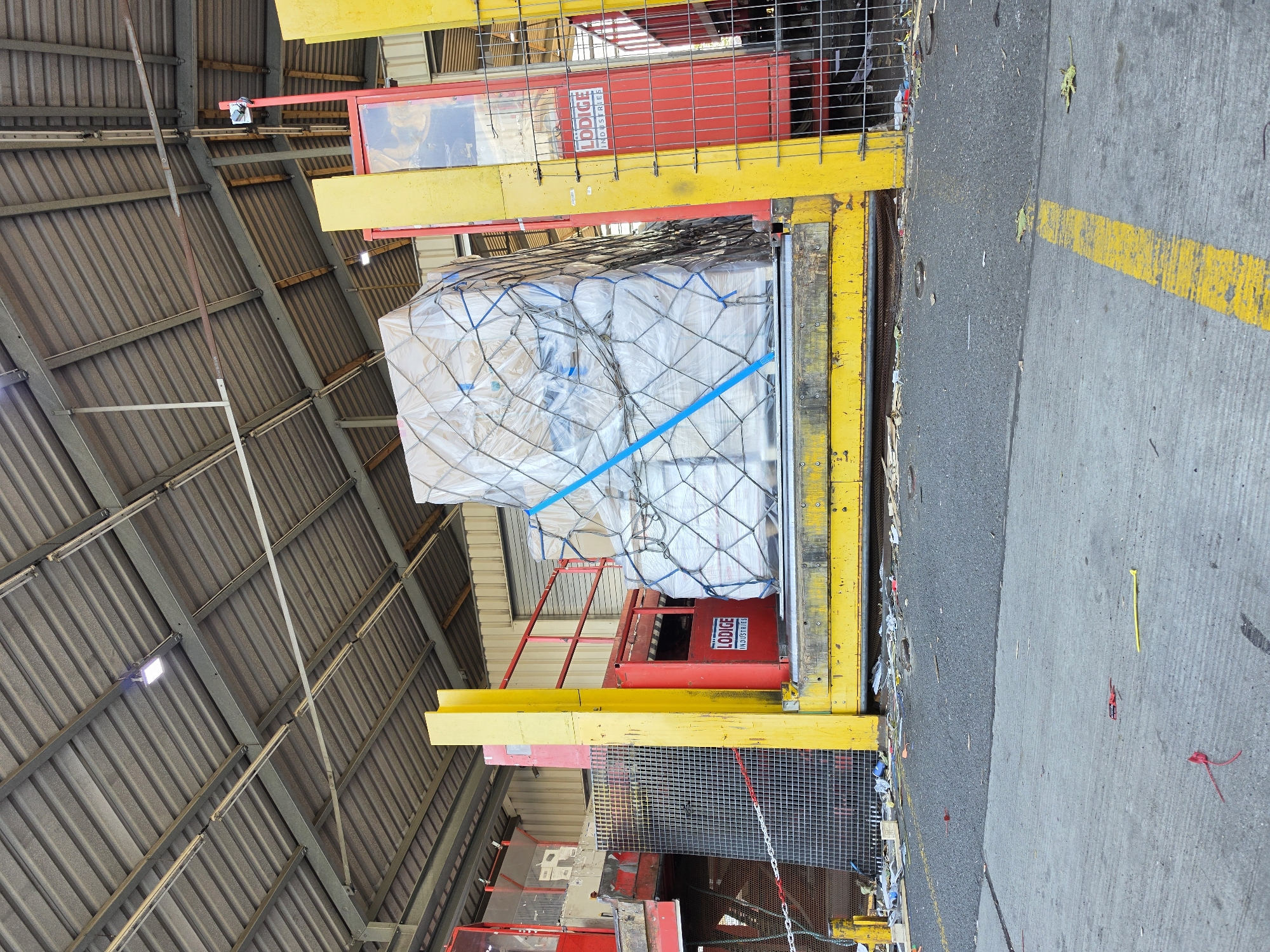 The image depicts a large, wrapped pallet of goods secured with netting and blue straps, positioned on a yellow loading dock platform. The platform is adjacent to a red and yellow structure, which appears to be part of a cargo handling system, possibly a lift or conveyor. A metal safety fence with a red railing encloses a portion of the area. In the background, the corrugated metal roof of a warehouse or industrial building is visible, supported by metal beams. Fluorescent lights are mounted on the ceiling. The ground in front of the loading dock is paved and shows scattered debris. On the red structure, a logo that reads "LODGE INDUSTRIES" is present. The scene suggests an active logistics or transportation hub. The location is identified as Frankfurt, Germany.