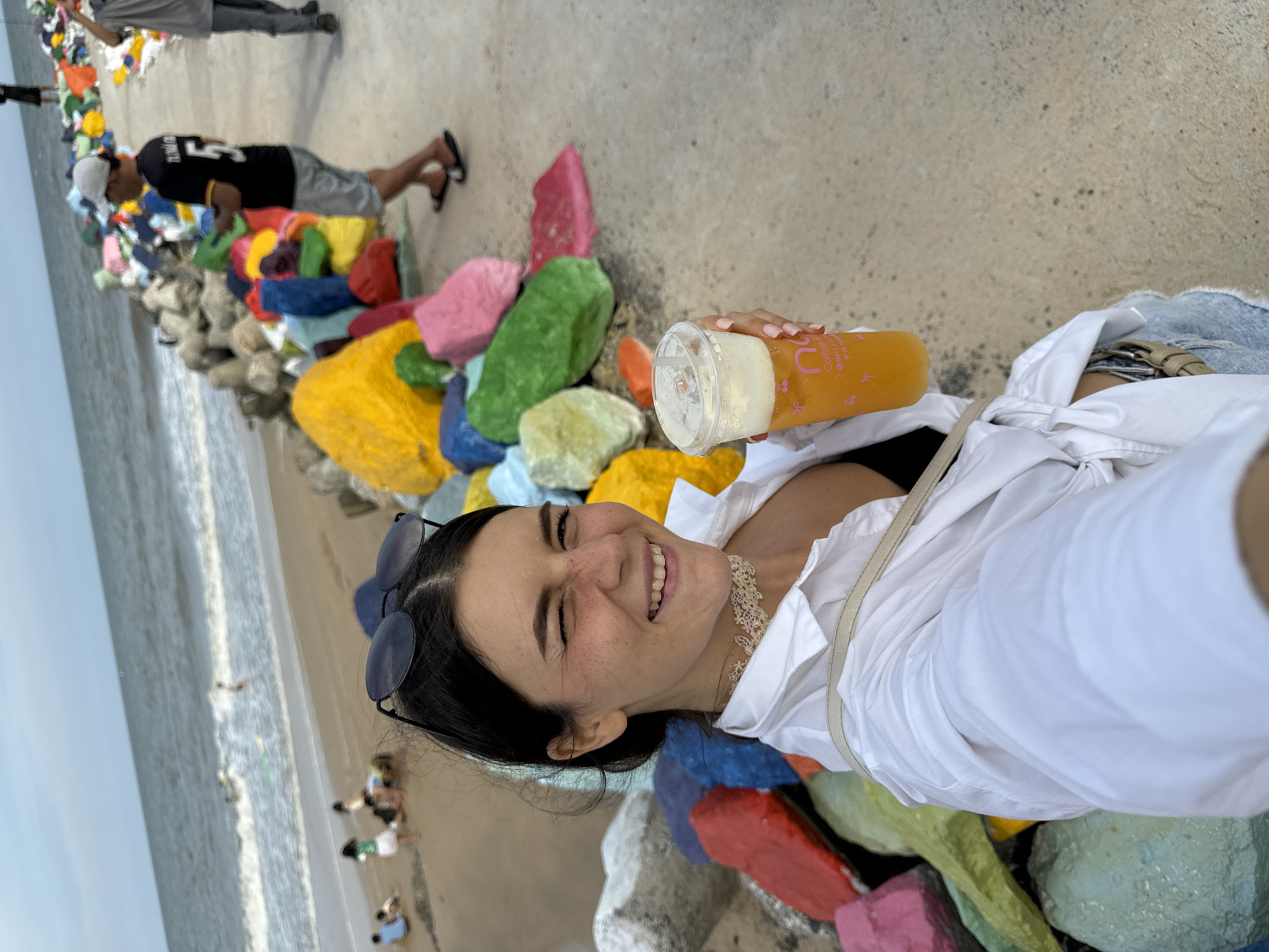 A young woman with dark hair and freckles is taking a selfie while holding a drink with a straw. She is wearing a white collared shirt and sunglasses perched on her head. She is smiling and winking at the camera. Behind her, a beach scene unfolds with the ocean, sand, and numerous brightly colored rocks. In the background, other people are visible on the beach and walking along a path. The colorful rocks appear to be part of a decorative installation along the waterfront, likely a tourist attraction in Mũi Né, Vietnam. The sky is overcast.