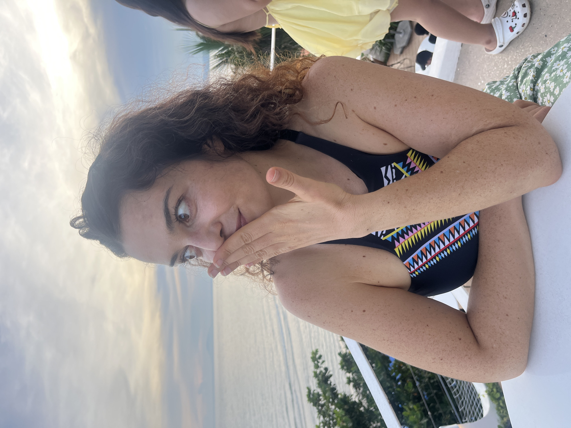 A woman with curly brown hair and fair skin is seated at a white table, with her left arm resting on the table and her right hand covering her mouth. She is wearing a black swimsuit with a colorful geometric pattern on the chest. Her eyes are wide and looking to her left. In the background, the sea is visible under a cloudy sky. To her right, a person wearing a yellow dress and white Crocs is partially visible, along with some greenery and a railing. The scene appears to be outdoors, possibly on a balcony or terrace overlooking the ocean. The location is identified as Mũi Né, Vietnam.