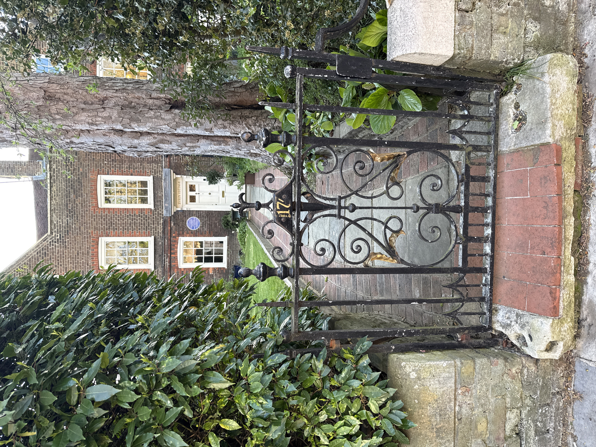 The image shows a decorative black iron gate at the entrance to Byron Cottage, located in the city of London, United Kingdom. The gate is situated between two stone pillars, opening onto a red brick pathway leading to the front of the cottage. The cottage is brick-built with white-framed windows and a white front door. A blue plaque is visible on the wall next to the door. A mature tree is behind the cottage. The scene is framed by greenery, with bushes on the left and plants visible through the gate. The photo is taken from a slightly low angle, looking towards the building.