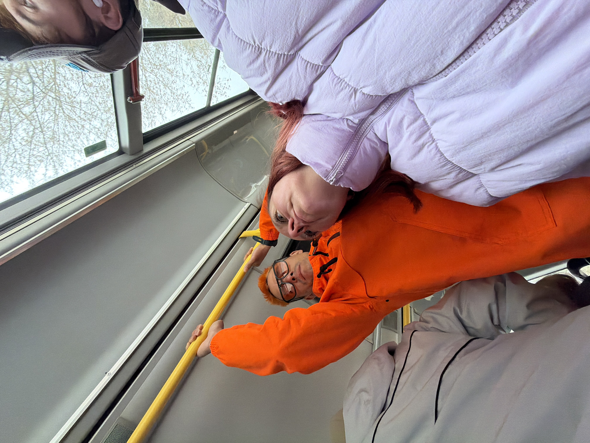 This image captures a moment inside a public transport vehicle, likely a bus or tram, in Chaikovsky, Russia. The photo is rotated 90 degrees clockwise, making the subjects appear upside down relative to the viewer.
Two individuals are prominently featured, looking directly at the camera. One person, positioned center-right, wears a vibrant orange jacket and glasses, and has reddish hair. They are holding onto a yellow handrail that spans the length of the vehicle's interior. A second person, situated slightly above and to the left of the first, is clad in a light lavender or purple puffer jacket, also with reddish hair visible. The top of a third person's head, wearing a grey baseball cap, is visible in the upper-left corner of the frame. Part of a fourth person, wearing a light grey jacket with black piping, is visible in the bottom-right.
The setting is characterized by the bright yellow handrails and the grey interior walls of the vehicle, typical of modern public transportation. Through the windows, bare tree branches are visible against a light sky, suggesting either late autumn, winter, or early spring. The bright natural light indicates it is daytime. The individuals appear calm, with their gazes directed towards the camera, possibly posing for a selfie or candid shot while commuting. No legible text is visible within the frame.