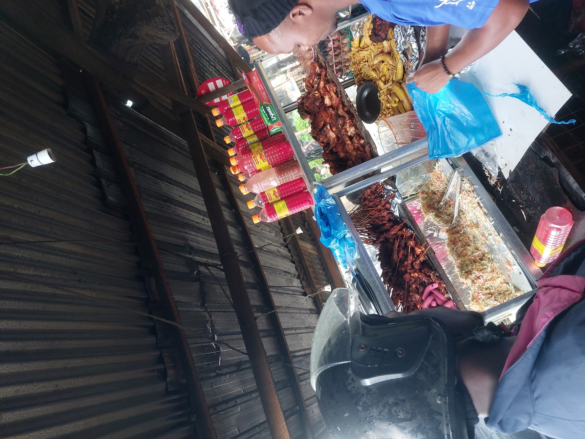 A street vendor in Dar es-Salaam, Tanzania, serves food from a metal stall. The stall contains fried plantains, grilled meat, and skewers. A person in a blue shirt is bagging the food. Bottles of red sauce are stacked along the side of the stall. The stall is located beneath a corrugated metal roof. A person with a helmet is present.