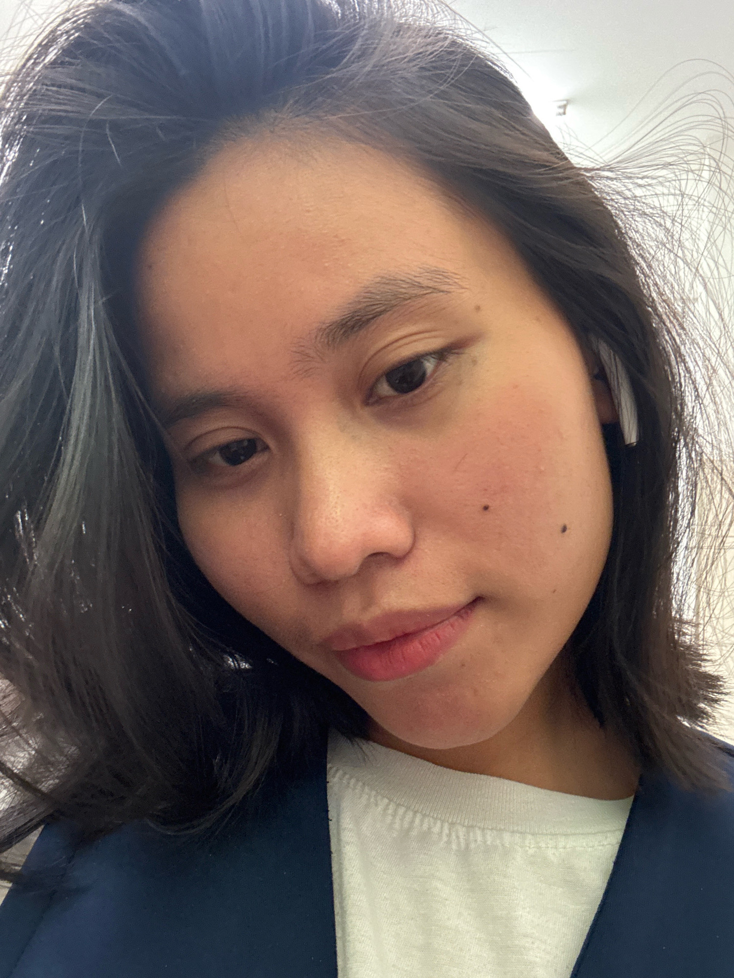 A close-up, front-facing portrait of a young woman with dark hair, taken indoors. She is wearing a white t-shirt underneath a dark blue jacket and an earpiece in her right ear. The background appears to be a white ceiling with a light fixture visible. Her expression is neutral, with her gaze directed slightly off-camera. The lighting suggests it's daytime, and the image is taken from chest level upwards. There are no explicit indicators of the city of East Jakarta, Indonesia, within the image itself.