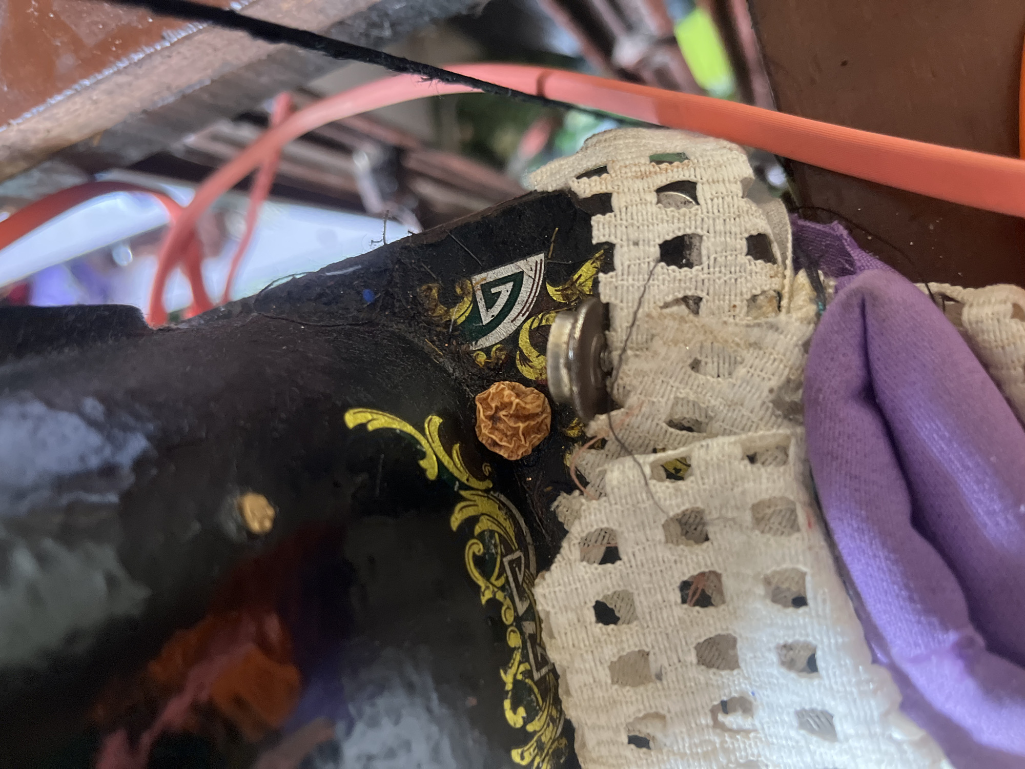 In Jauro Yinu, Nigeria, a close-up, eye-level shot captures a detail of an antique sewing machine. The machine's black enamel surface is adorned with yellow floral decals and a small triangular emblem. A small, dried orange object, possibly a seed or dried berry, rests on the machine's surface near a silver screw. A white lace trim is attached to the machine, with a purple fabric bundle visible beneath it. The background is blurred, suggesting an indoor or workshop setting, with hints of other objects and materials, including an orange cord.