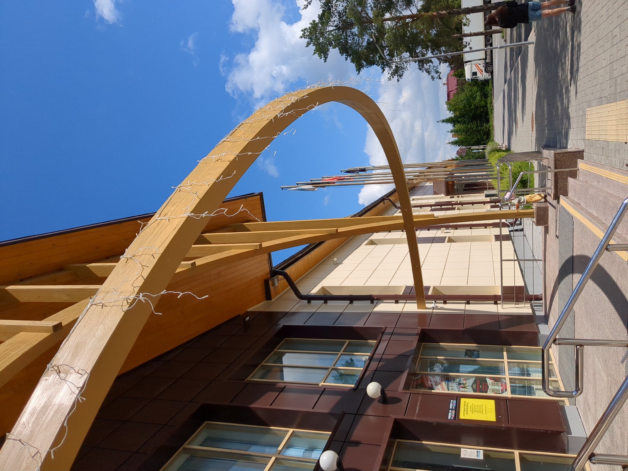 This image depicts the exterior of the "Neftyanik" Sports Complex, located at Ulitsa Osipenko, 83, in Tyumen, Russia, under clear skies. The building features a modern design with a facade composed of dark brown and light beige rectangular panels, punctuated by multiple large windows with light-colored frames. A prominent, curved wooden beam, part of an architectural canopy or entrance feature, extends from the building's upper structure. This beam is strung with numerous white, bare-bulb string lights.
The sky is a vivid blue with scattered white cumulus clouds, indicating bright daylight and strong sunlight, which casts distinct shadows. In the foreground, a set of concrete steps with polished metal railings leads to the building's entrance. Yellow tactile paving is visible on the ground before the steps. A yellow sign on the glass entrance door reads "СПОРТИВНЫЙ КОМПЛЕКС "НЕФТЯНИК"" (Sports Complex "Neftyanik") along with other Cyrillic text and QR codes.
To the right of the entrance, a paved walkway extends into the distance. A person is visible on this walkway, standing with their back to the viewer, wearing blue shorts and a dark top. A small, partially obscured figure, possibly a child, is near the entrance steps to the left. Beyond the paved area, a road is present, where a white semi-truck with a trailer is visible. Several tall, green pine trees are situated to the right of the walkway and road, and other green foliage and distant structures are visible. A series of flags on poles stand near the building's entrance, below the wooden arch.