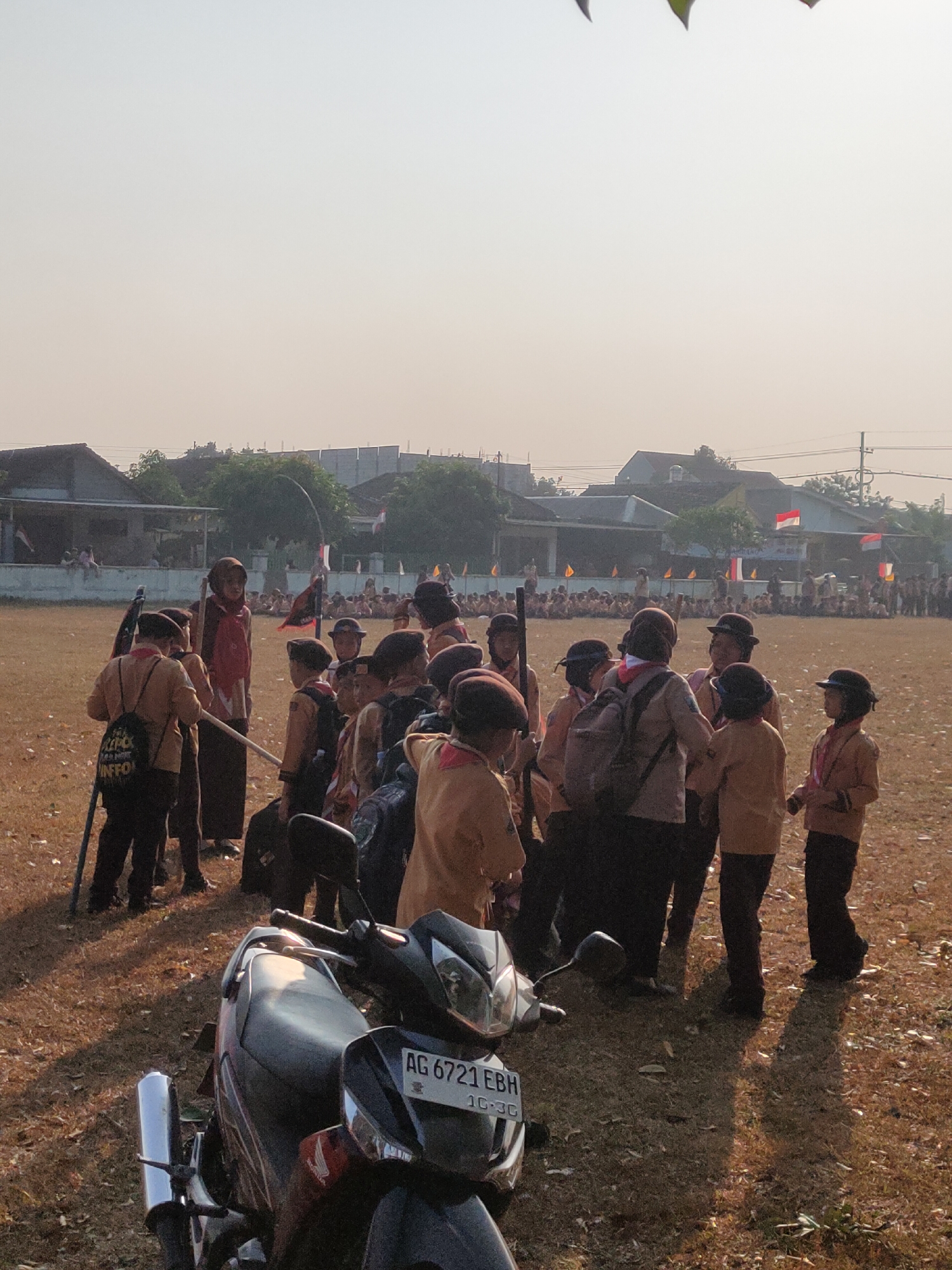 This is a medium shot of a group of young people dressed in scout uniforms, gathered in an outdoor grassy area under a hazy sky. The people are predominantly wearing brown and red uniforms, with some wearing hats. Several individuals are holding sticks or poles. In the foreground, a motorcycle with the license plate AG 6721 EBH is parked. In the background, there are buildings with tiled roofs, trees, and flags, including what appear to be Indonesian flags. The ground is covered in dry grass and fallen leaves. The scene suggests an outdoor gathering or event, possibly a scout jamboree or ceremony, taking place in a semi-urban or rural setting in Ngasem, Indonesia.