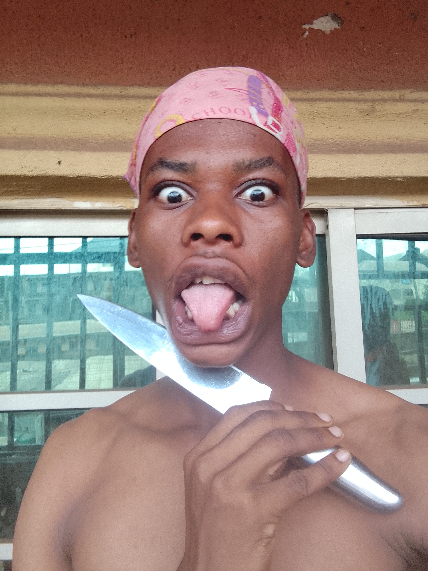 A shirtless person with dark skin, wearing a pink headscarf, holds a silver knife against their neck. The person has wide eyes and their tongue is sticking out of their mouth. The location appears to be indoors, in front of a window. The location is in the city of Unknown, Unknown.