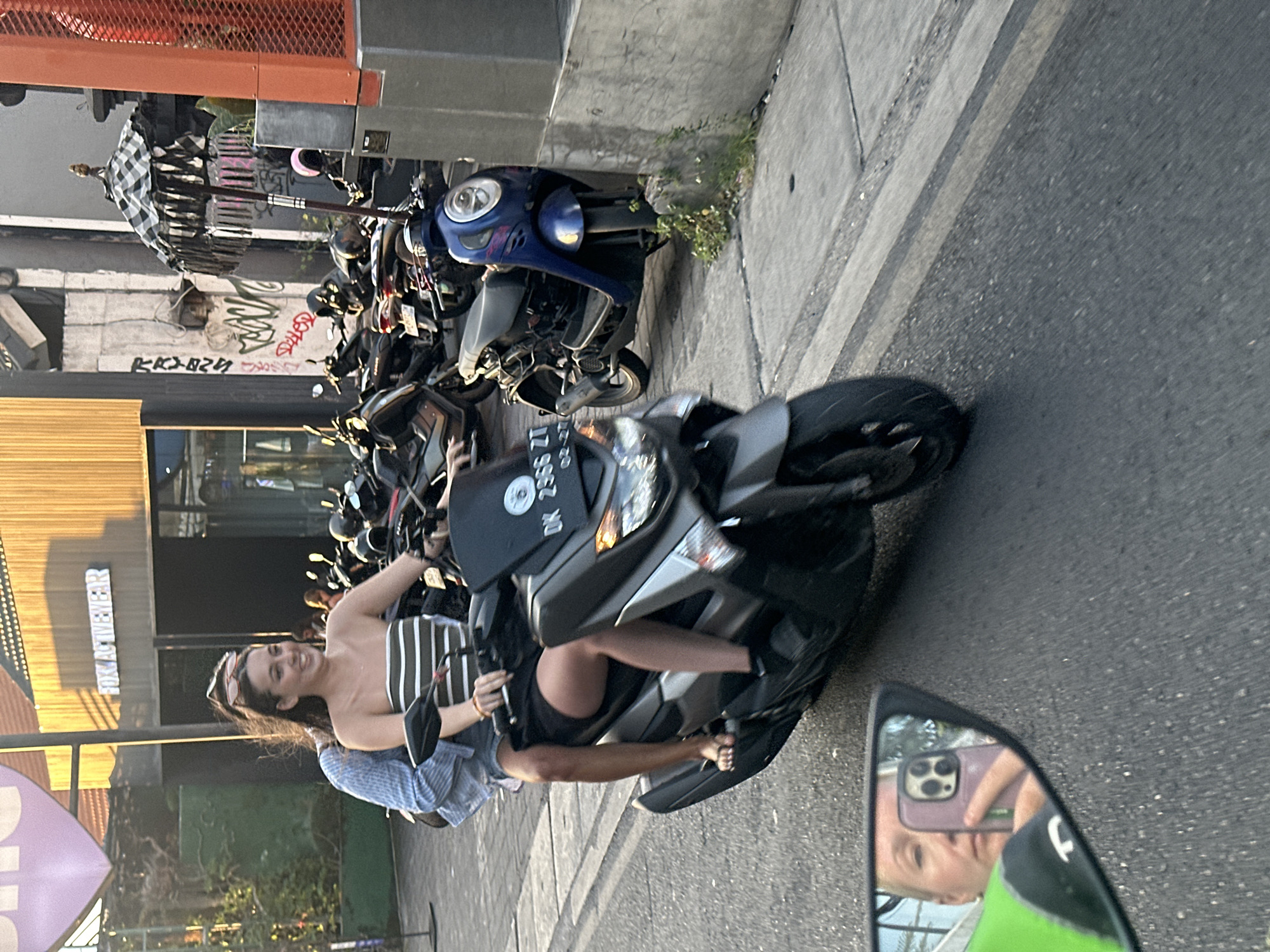 Two individuals, a male driver and a female passenger, are riding a dark gray scooter in the city of North Kuta, Indonesia. The female passenger is wearing a strapless, horizontally striped top and dark shorts, with her legs visible. The male driver is wearing a blue and white striped long-sleeved shirt. The scooter has a license plate that reads "DK 2366 ZT". In the foreground, a motorcycle's mirror reflects a woman holding a purple smartphone, taking a selfie. The background features buildings with signage, including "FOX ACTIVEWEAR", and numerous parked scooters and motorcycles. The scene appears to be on a street with a sidewalk and indicates a daytime setting with warm lighting.
