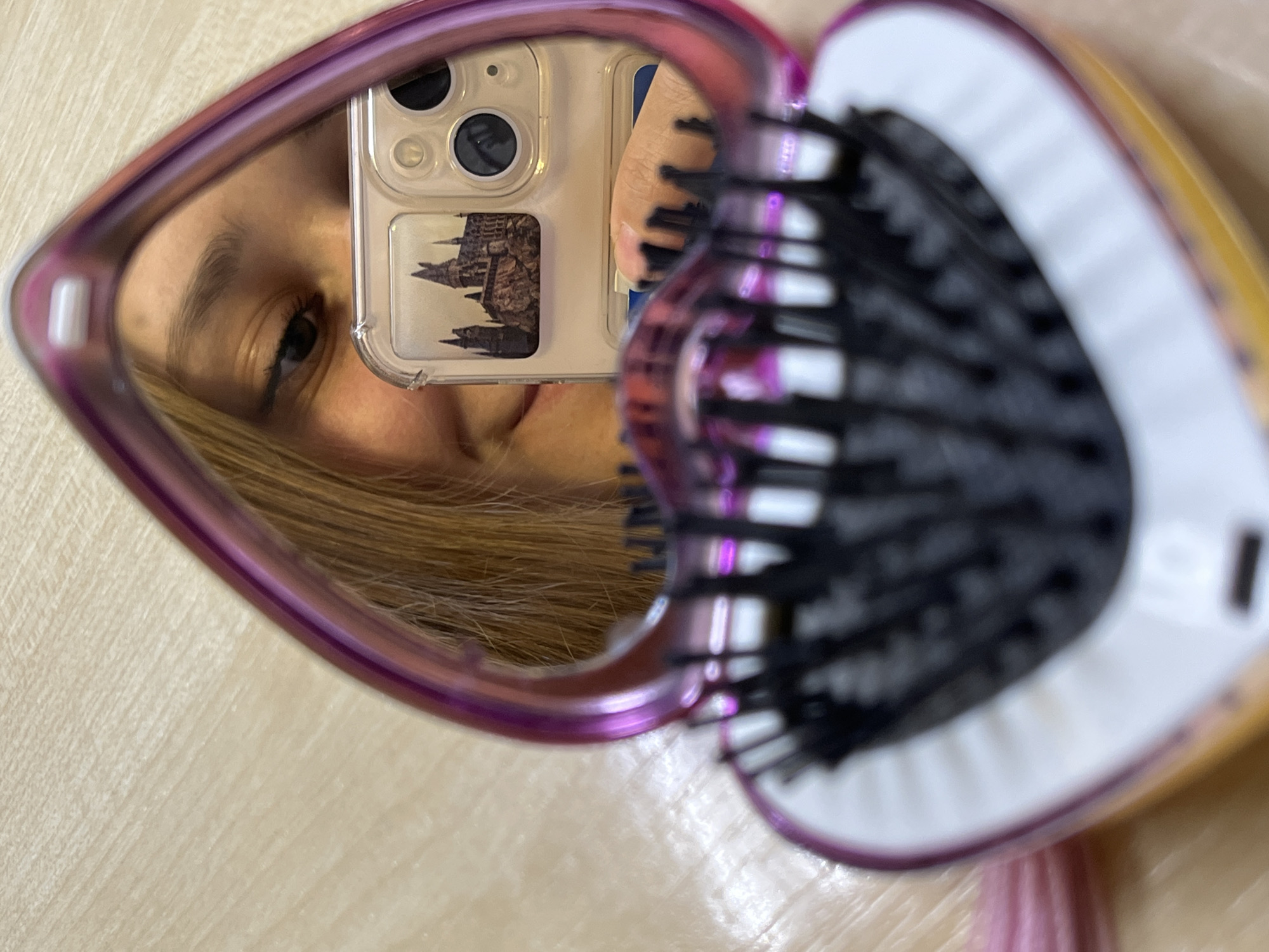 A person is reflected in a heart-shaped mirror next to a hairbrush. The reflection shows part of a face, blonde hair, and a cell phone in a clear case with a building design on the back. The hairbrush has black bristles. The surface below the mirror is light colored. No location information can be derived from the image.