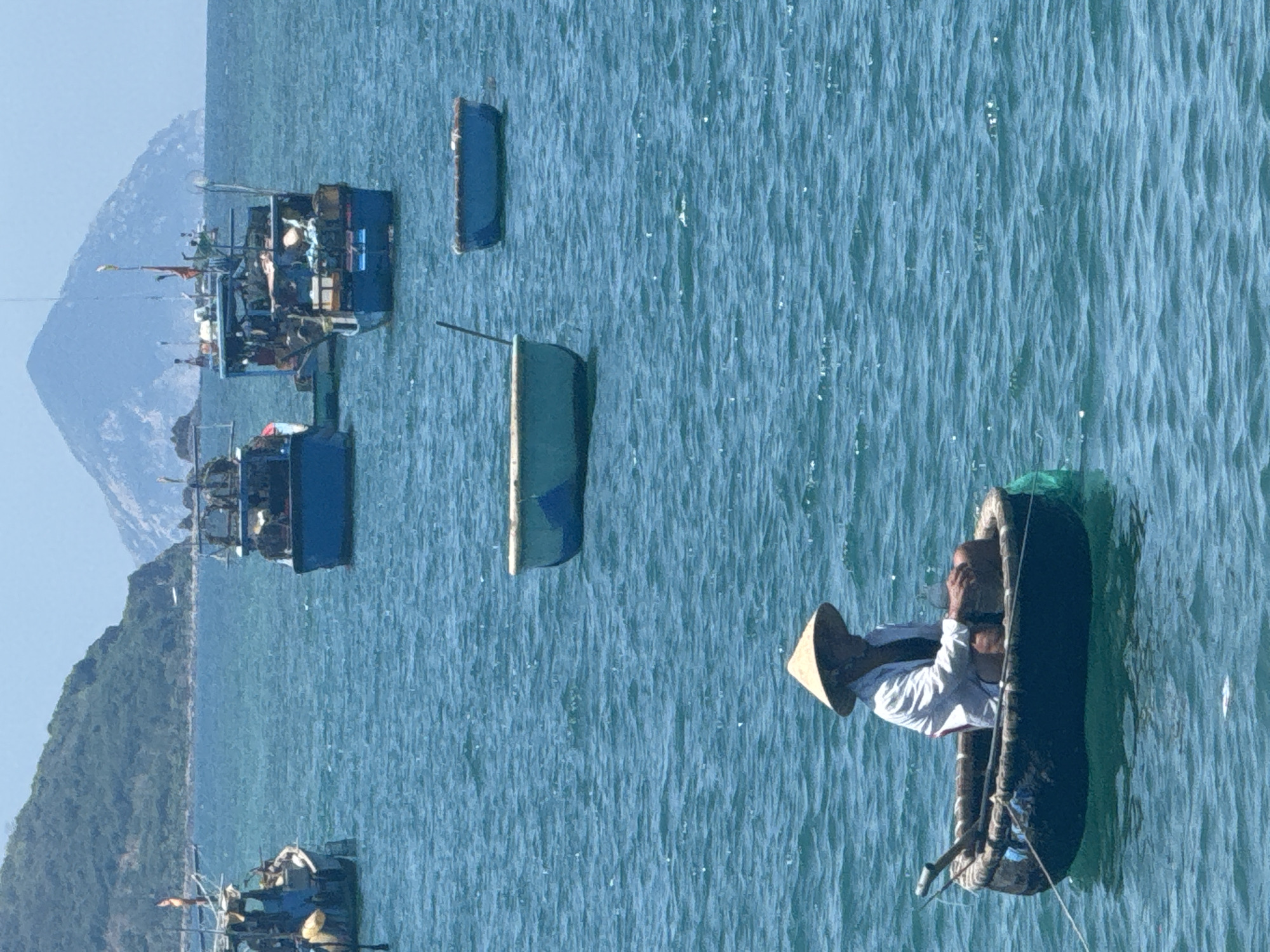 An overhead view captures a tranquil blue ocean dotted with various boats. In the foreground, a solitary figure wearing a conical hat sits in a round, dark boat, appearing to be fishing. Further back, larger blue fishing vessels are anchored, their decks laden with equipment. In the distance, a lush green island rises from the water, topped by a hazy, distant mountain peak under a clear sky. The water is slightly choppy, with sunlight reflecting off its surface.