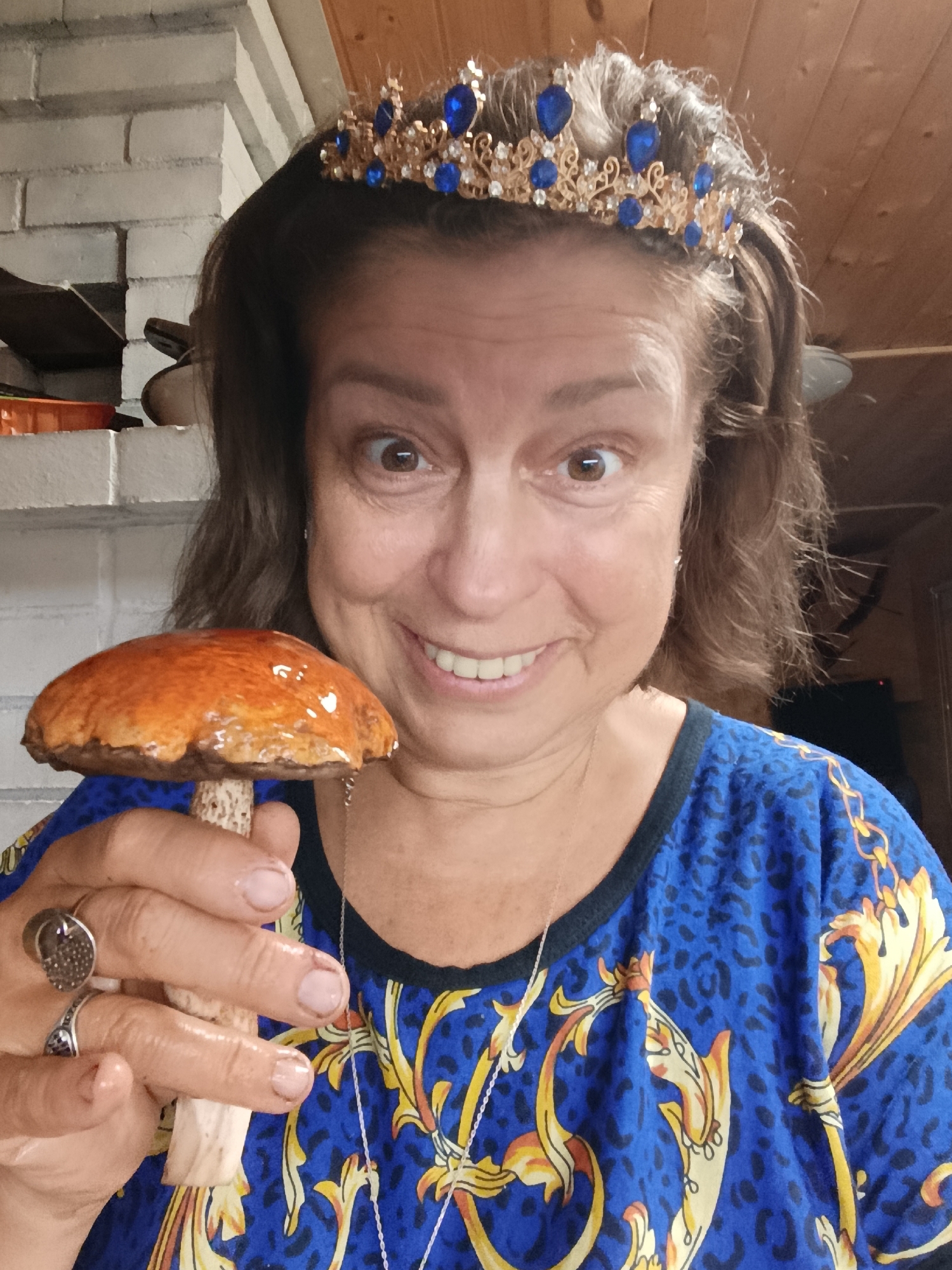 In a medium shot, a woman with brown hair and a tiara on her head is smiling at the camera while holding a mushroom. She is wearing a blue shirt with a leopard print and gold chain pattern. The background appears to be indoors, with a white brick wall and wooden paneling visible. The woman's left hand holds the stem of the mushroom, and her right hand is near her chest. The mushroom has a reddish-brown cap and a light-colored stem.
