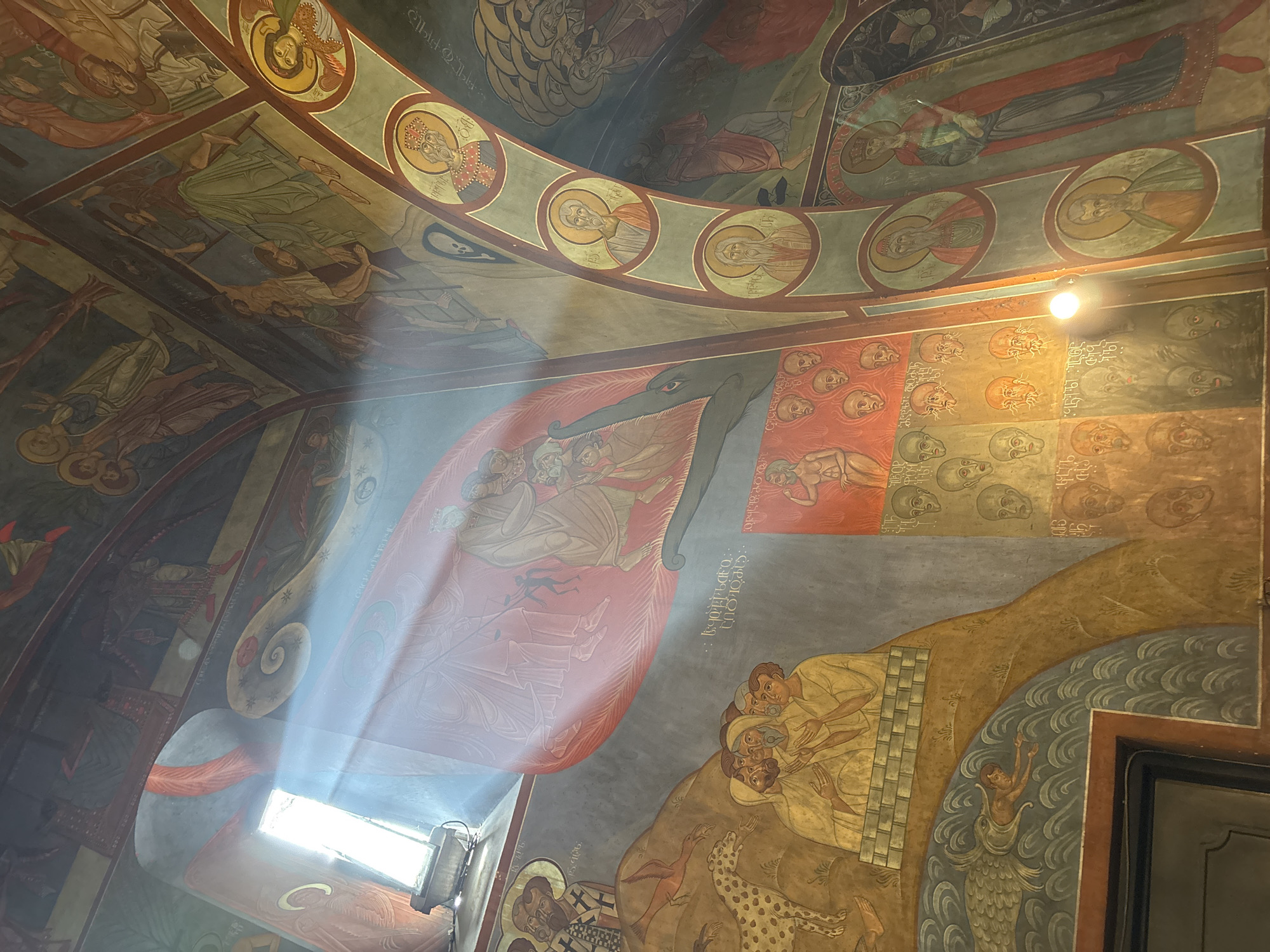 The image shows an interior view of a room adorned with religious frescoes. A strong beam of sunlight streams in from a window, illuminating the murals on the walls and ceiling. The frescoes depict various scenes, including figures resembling saints and biblical narratives. In the lower right portion, there are depictions of people, animals, and what appears to be a mermaid figure. In the center, there is a red-hued fresco showing people being dragged, with a line of faces overhead and text in an unknown language. The image is taken at Tbilisi, Georgia, specifically inside the Anchiskhati Basilica.